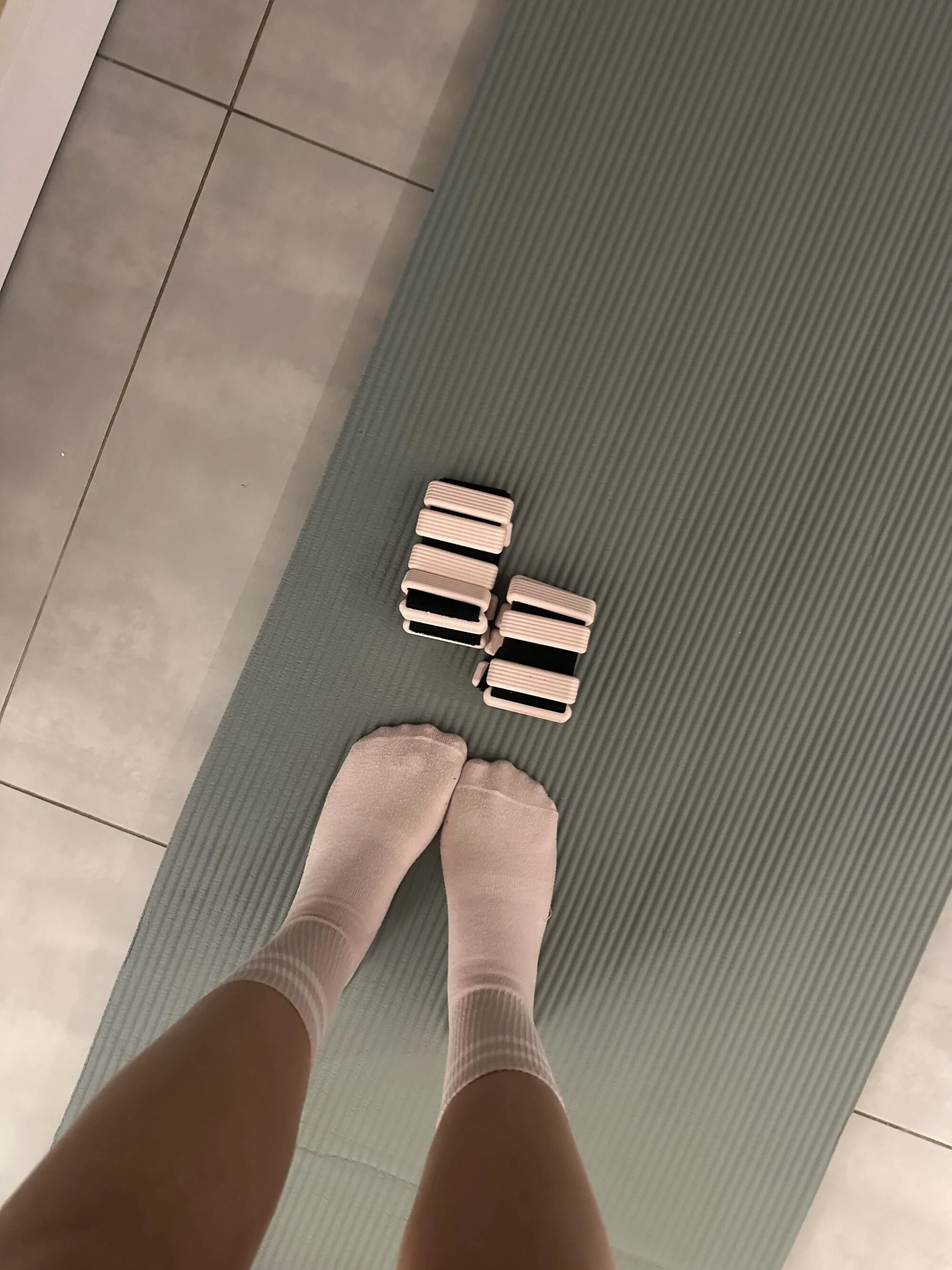 A person's feet in light pink socks stand on a green yoga mat, with several small, rectangular, pink and black objects arranged nearby.