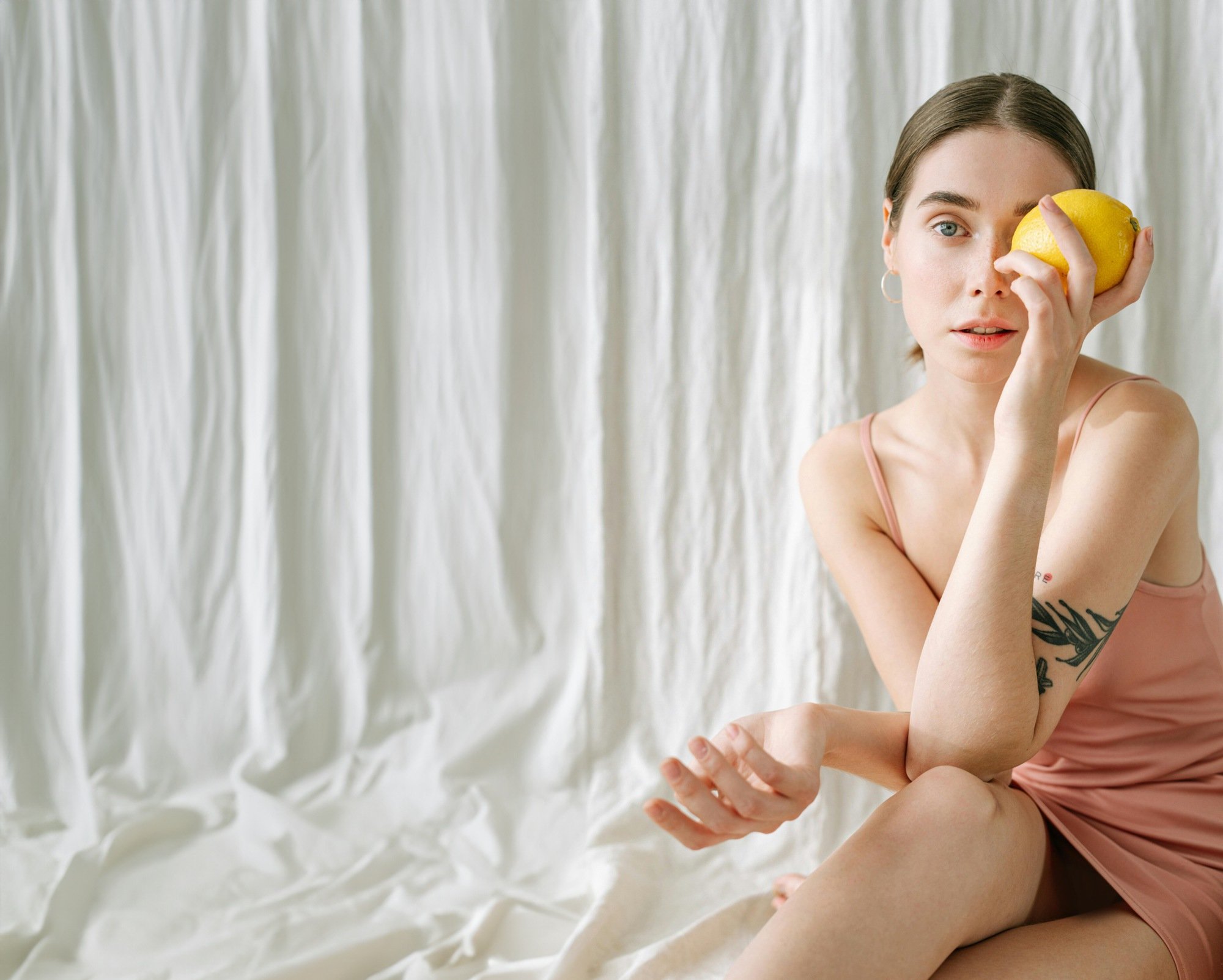 A young woman with short brown hair and a tattoo on her arm is sitting in front of a cream-colored curtain, holding a lemon near her eye and looking at the camera, wearing a pink sleeveless dress.