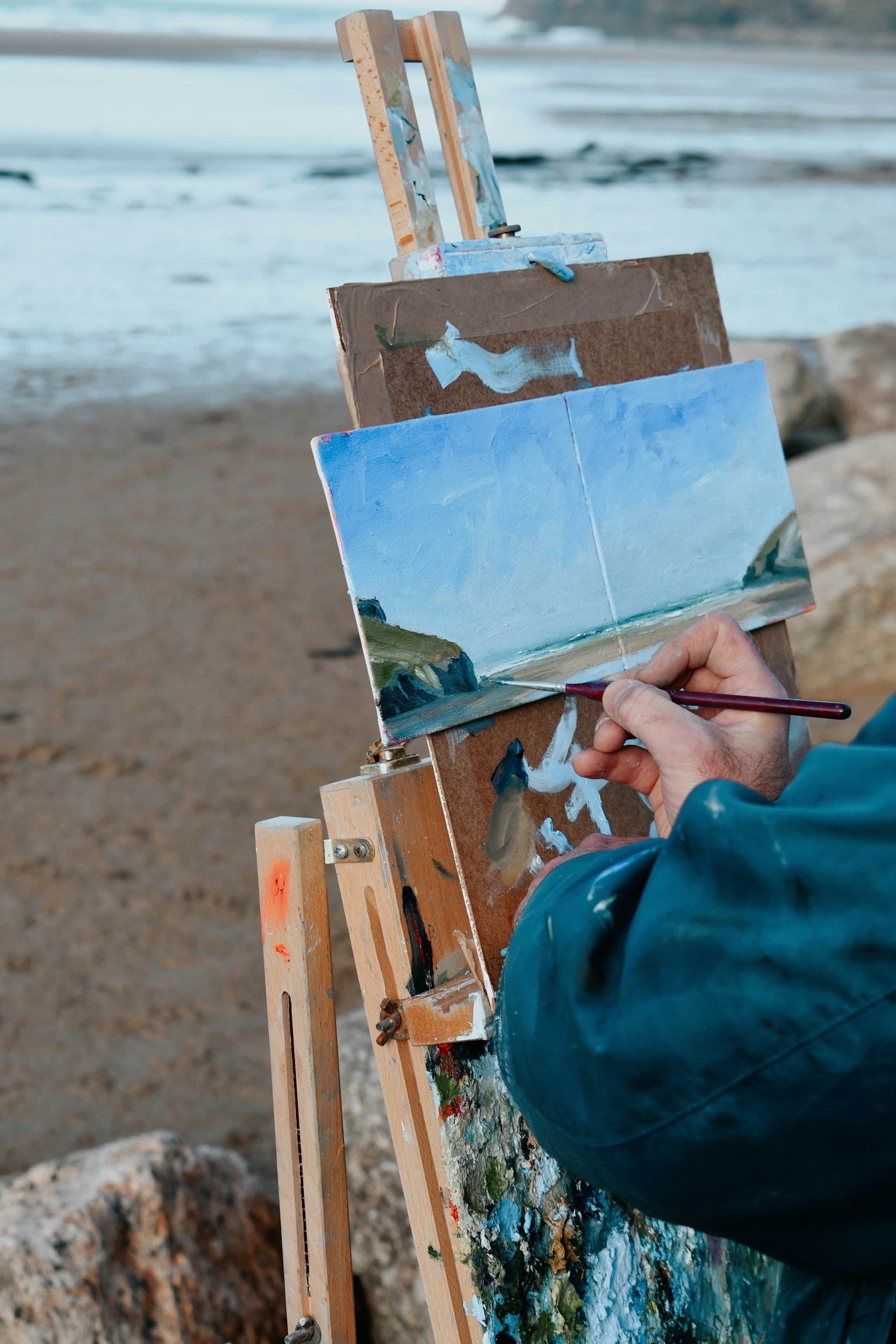 Person painting a seascape on a small canvas outdoors near a beach, using an easel and a paintbrush.