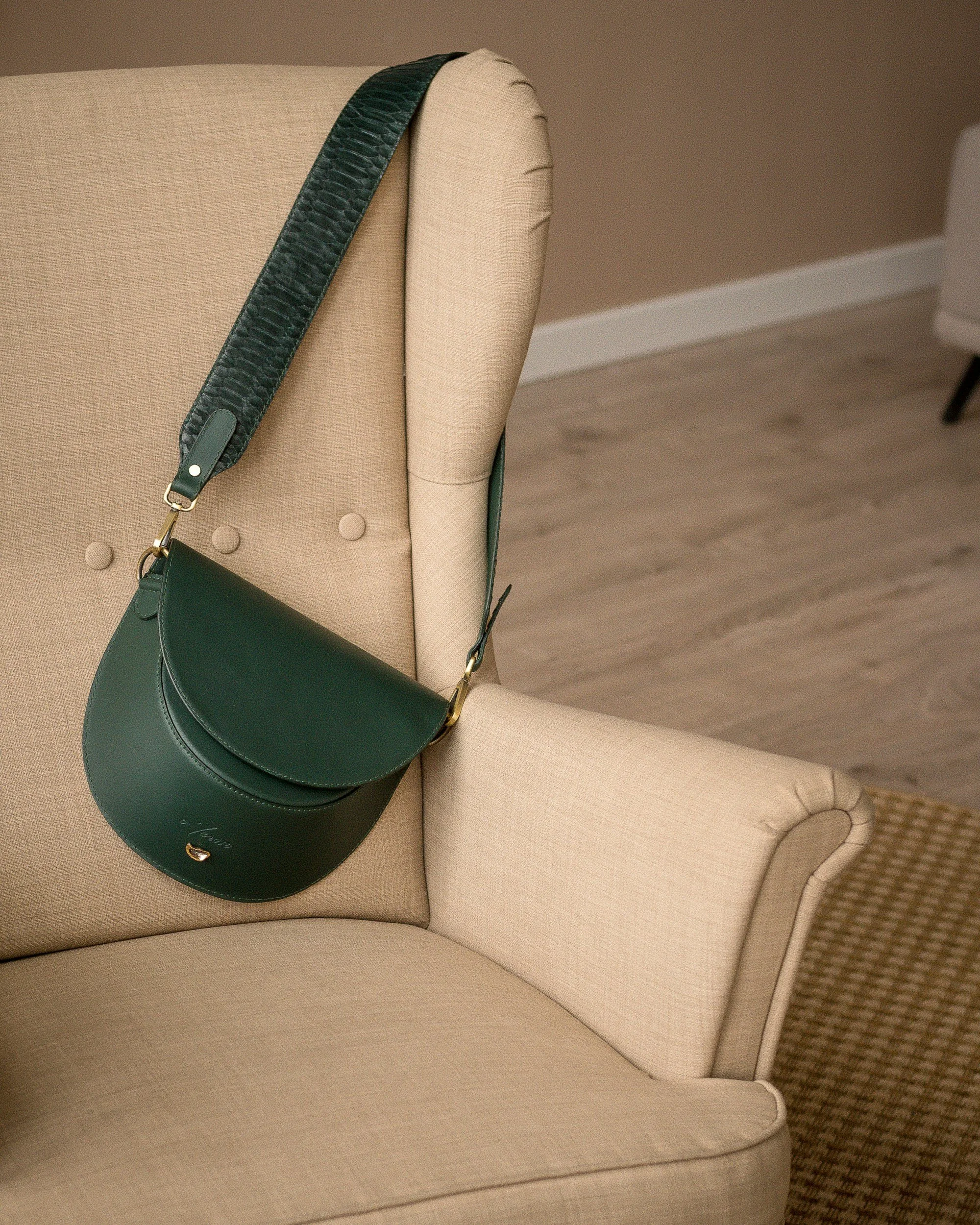 Green leather handbag hanging on the back of a beige upholstered armchair.