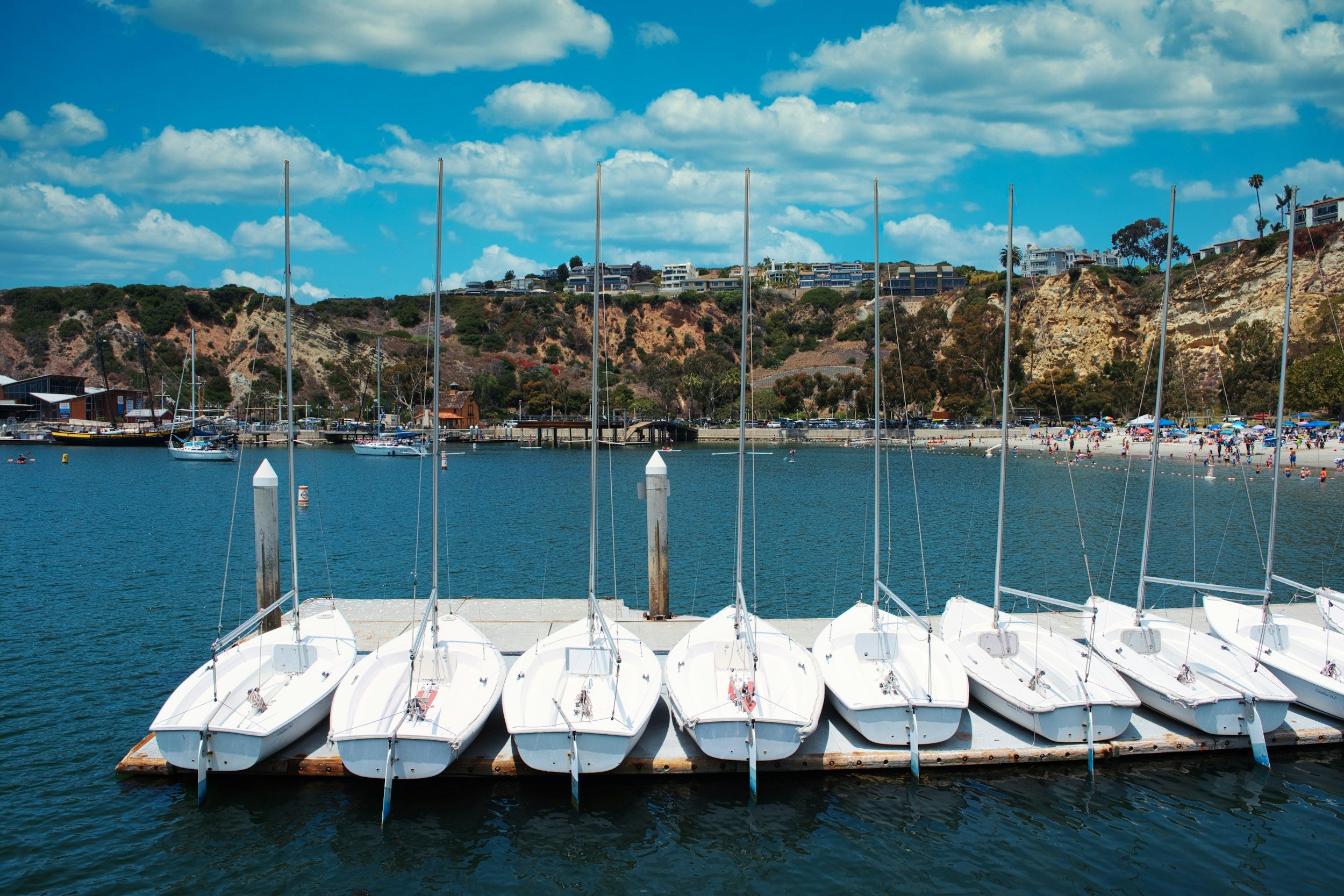 A marina with seven white sailboats docked side by side on calm blue water, with a sandy beach and hillside in the background under a partly cloudy sky.