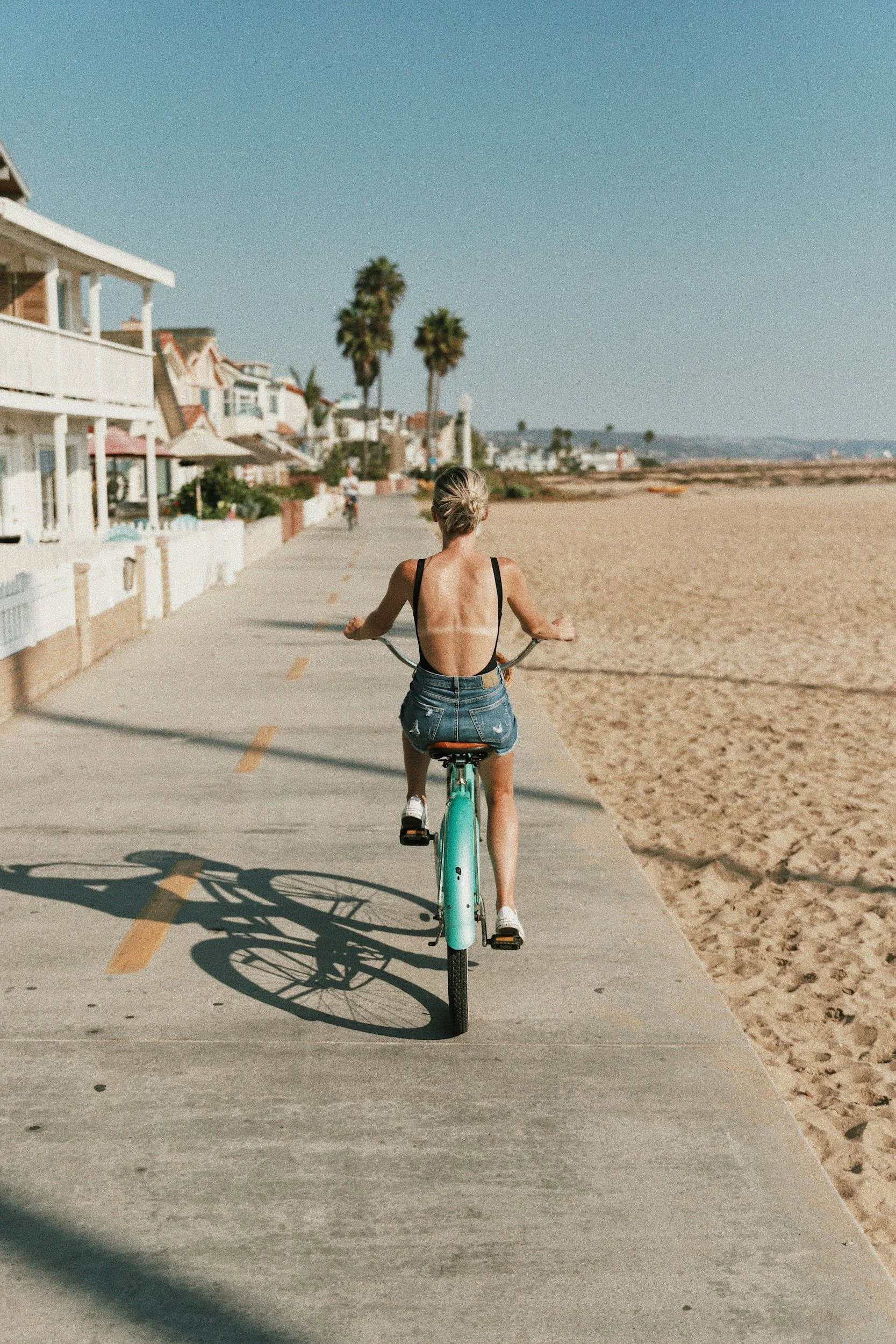 A woman with short blonde hair riding a mint green bicycle in Newport Beach, wearing a backless top and denim shorts under a clear sky.