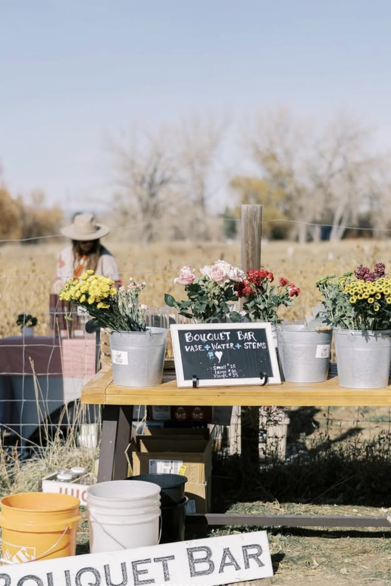 A small bouquet bar stand with buckets of colorful flowers, including yellow, pink, and purple, in a farm or outdoor setting with a woman in a wide-brimmed hat in the background.