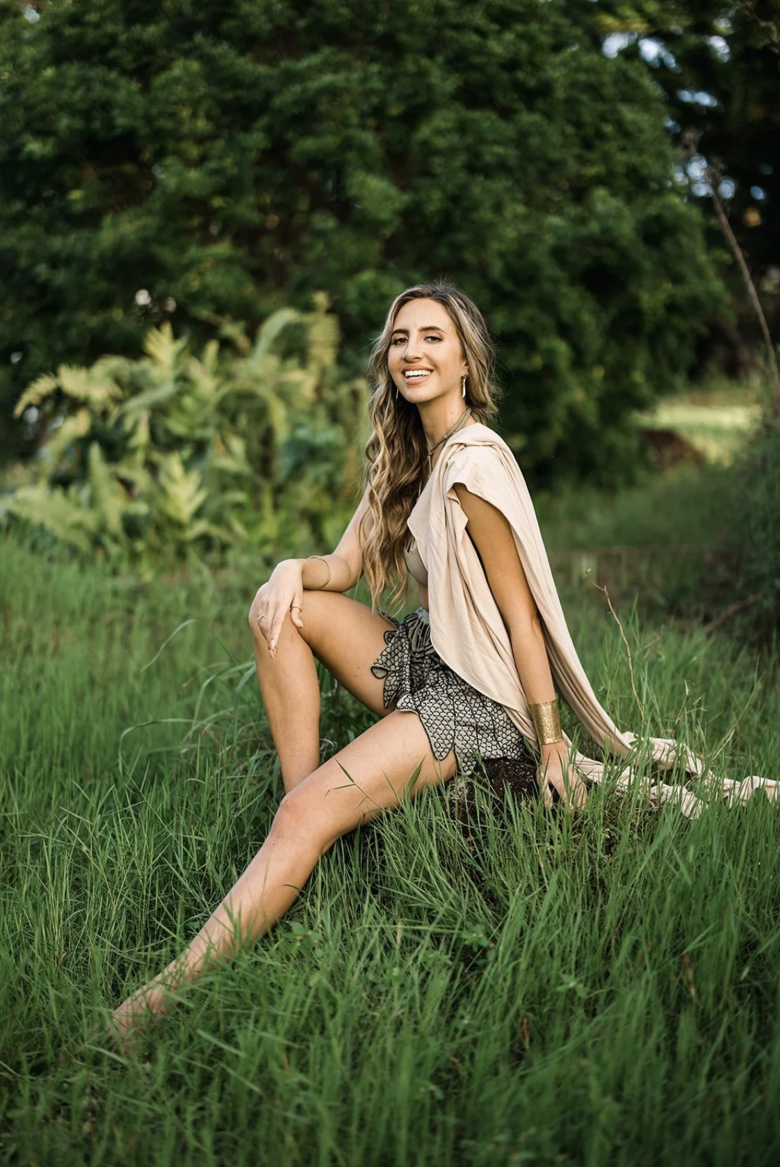 A woman with long wavy hair sitting outdoors on green grass, smiling, with a lush background of trees and plants.
