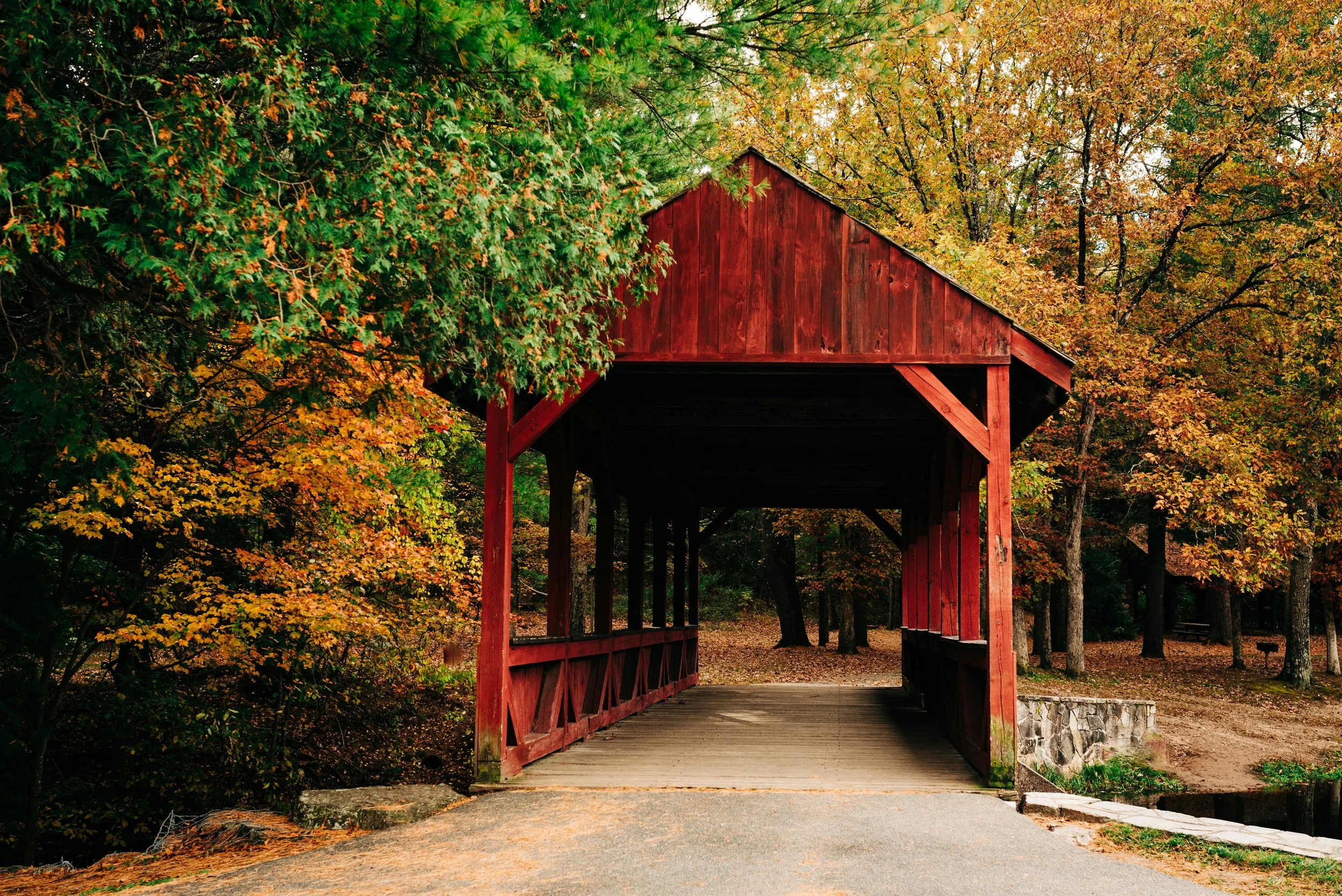 A red covered bridge surrounded by colorful autumn trees with orange, yellow, and green leaves in a park setting.