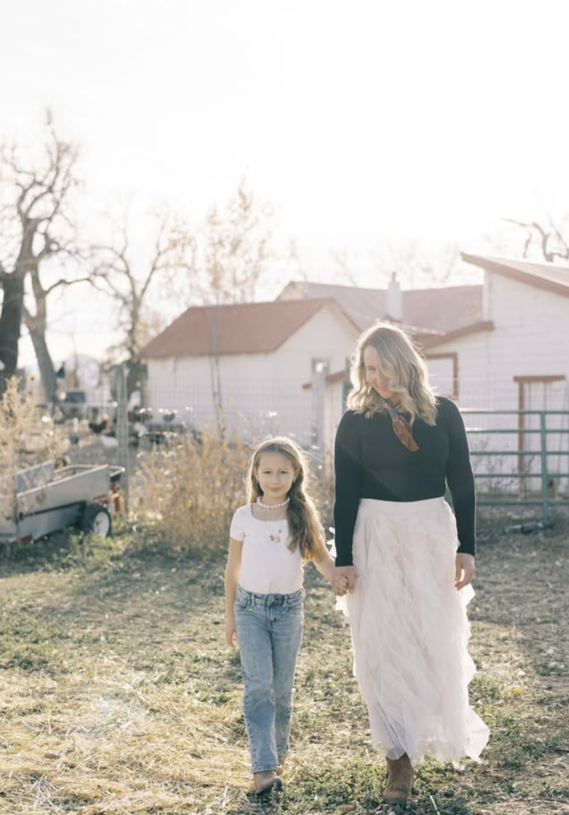 A woman and a young girl walking hand in hand in a farm or rural backyard on a sunny day.
