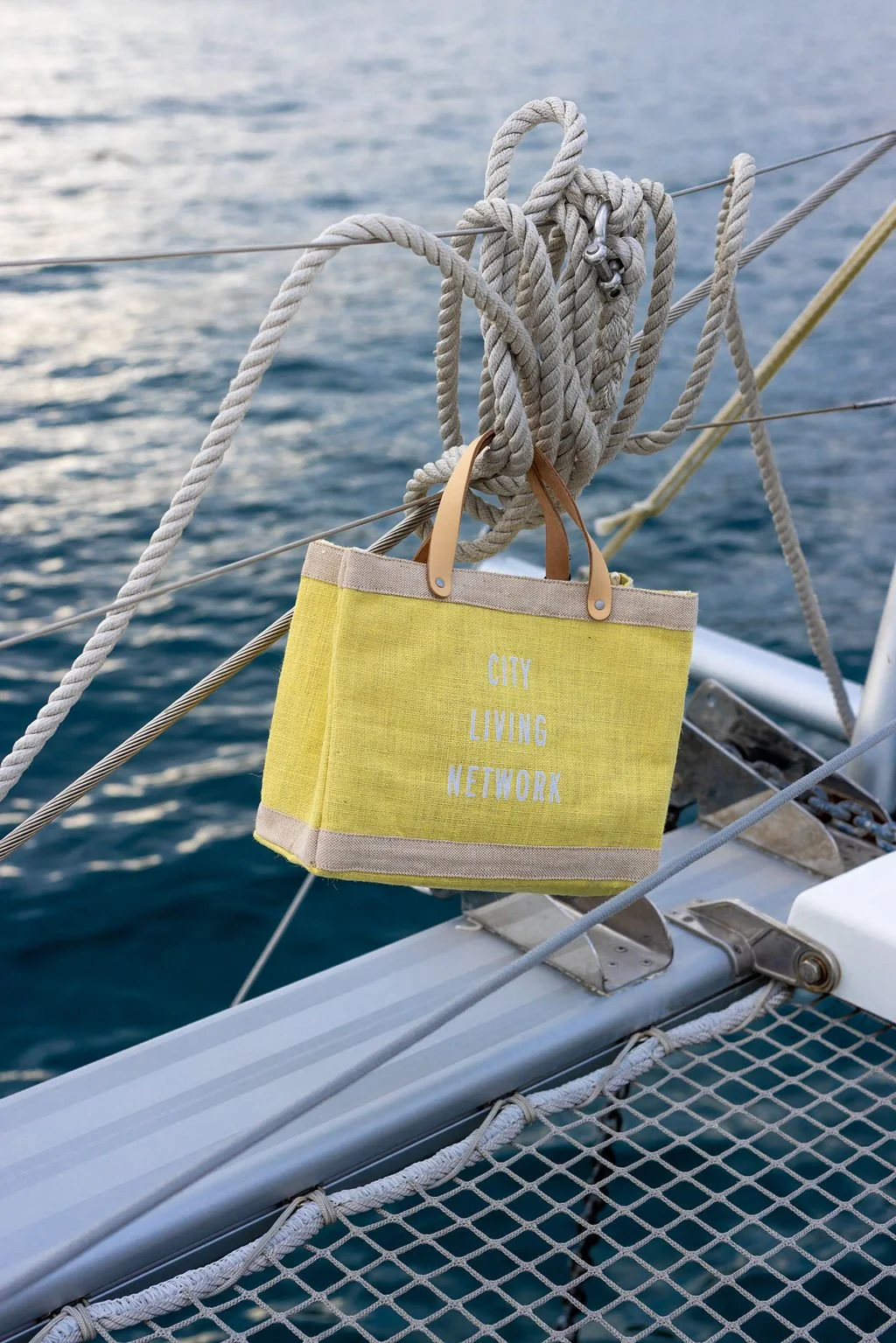 A yellow tote bag with brown handles hanging on a rope on a boat, with a calm ocean in the background.