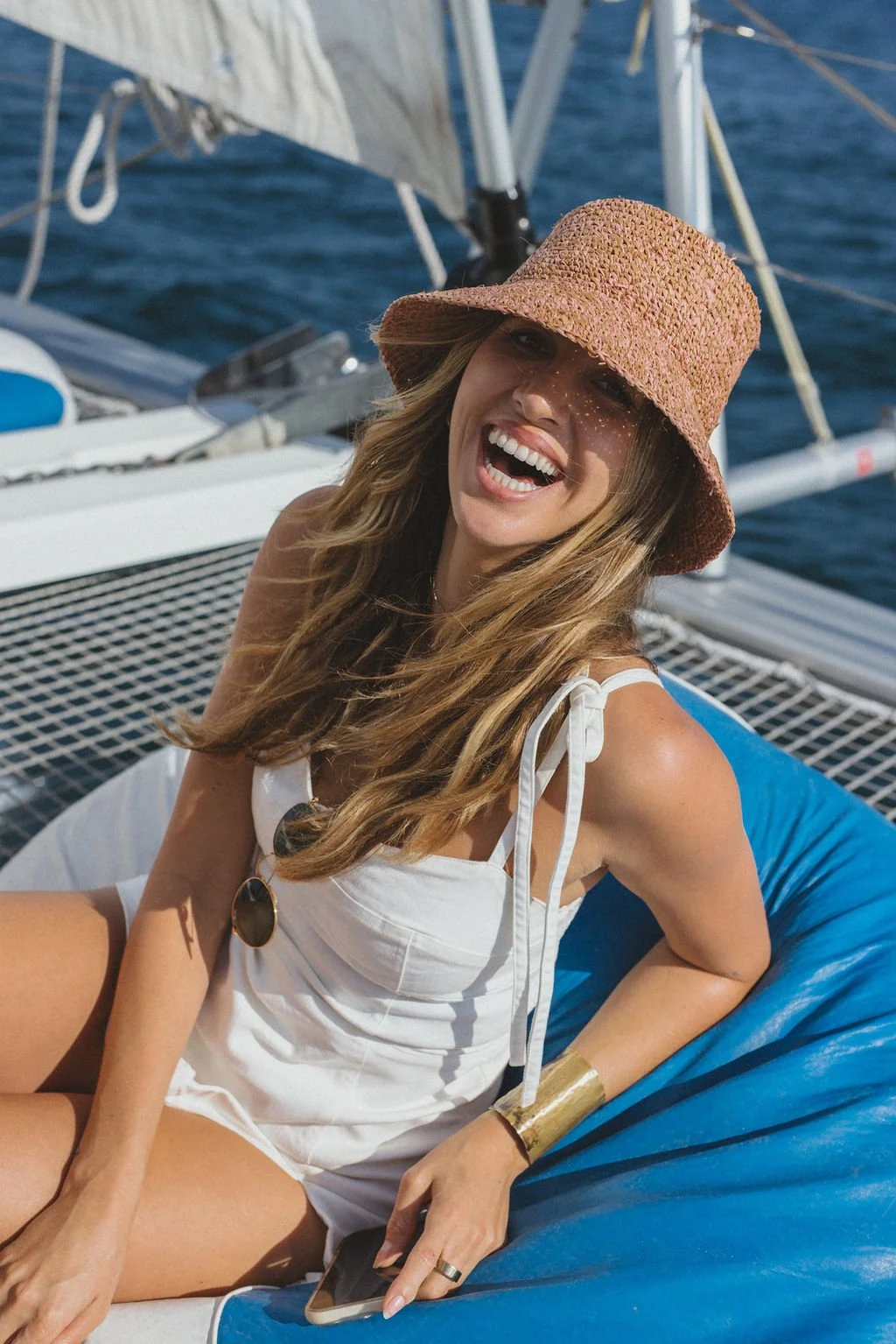 A smiling woman wearing a straw hat and white outfit sitting on a blue bean bag on a boat, with water and boat rigging in the background.
