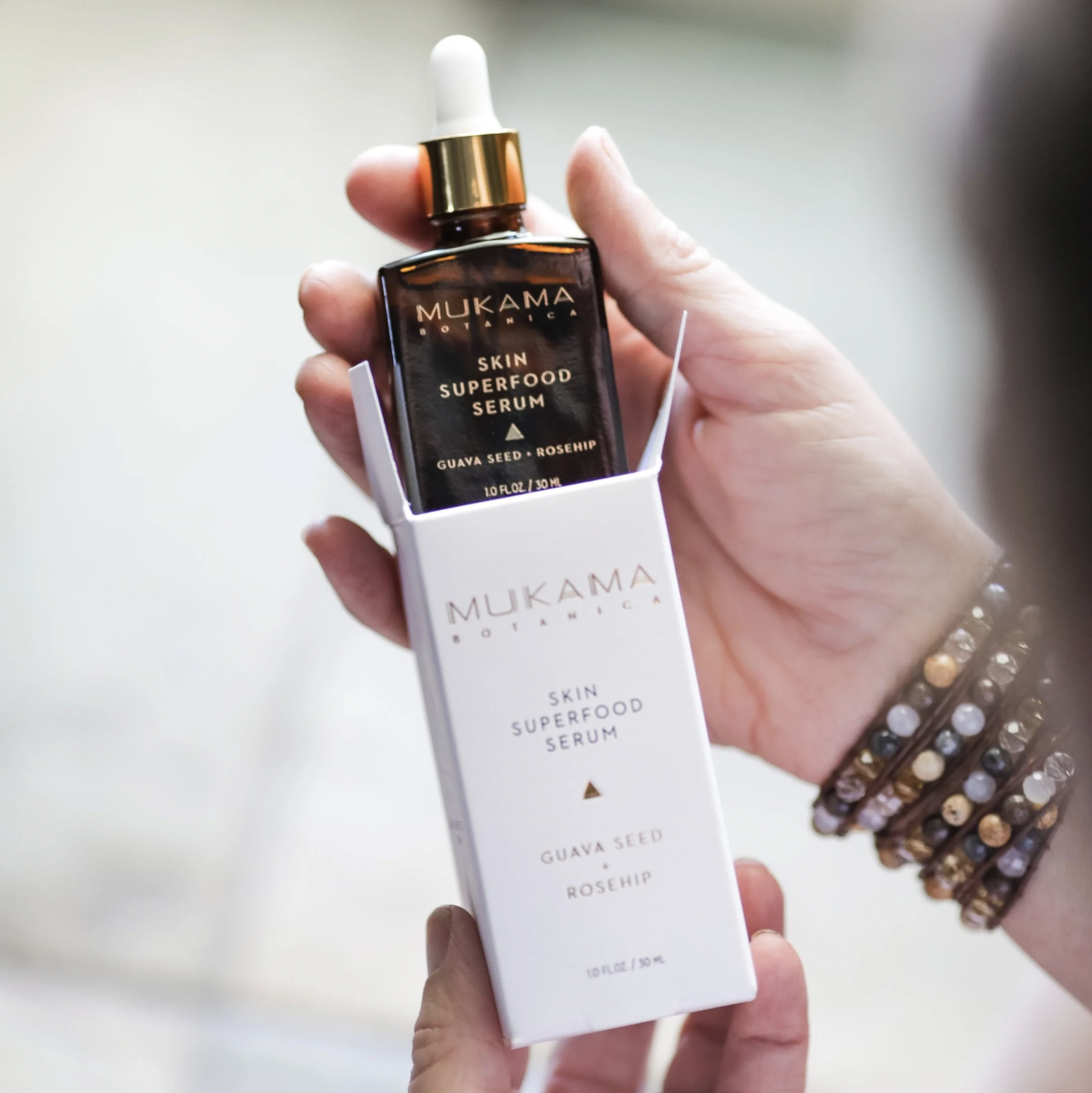 A hand holding a brown glass bottle of Mukama Botanica Skin Superfood Serum with a white dropper cap. The bottle is partially inside a white box displaying the product's name and ingredients, including guava seed and rosehip.
