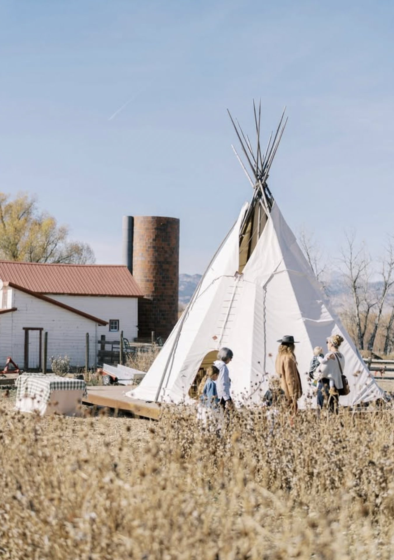 People gathered around a large white teepee in a field during daytime with clear skies and some trees in the background.
