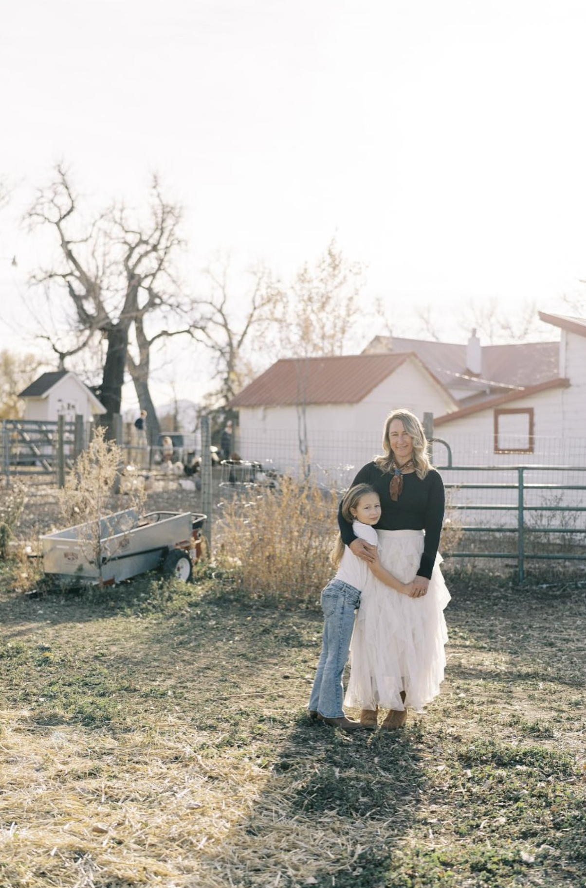 A woman and a young girl hugging and smiling on a farm with trees, a shed, and a trailer in the background during daytime.