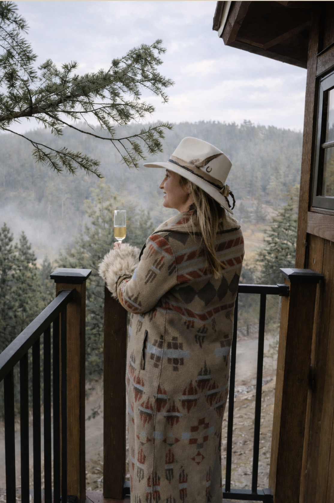 A woman standing on a wooden balcony overlooking a forest, holding a glass of white wine, dressed in a patterned coat and wide-brimmed hat.