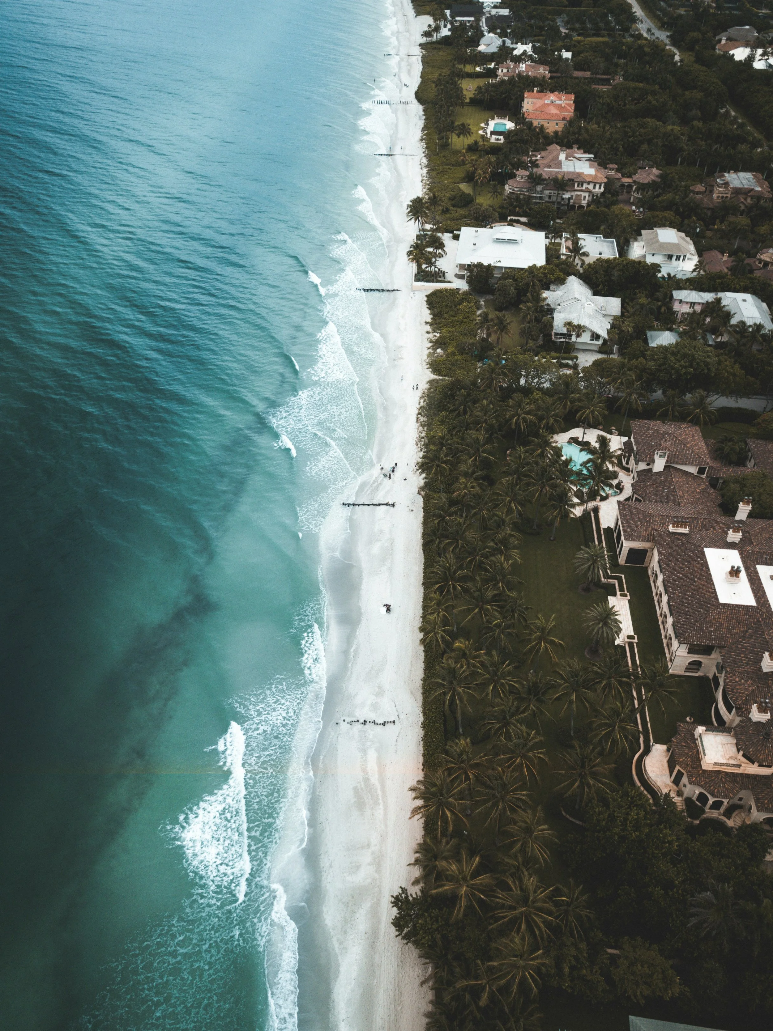 Aerial view of a beach with turquoise water, white sand, and waves, bordered by lush green trees and residential houses with pools.