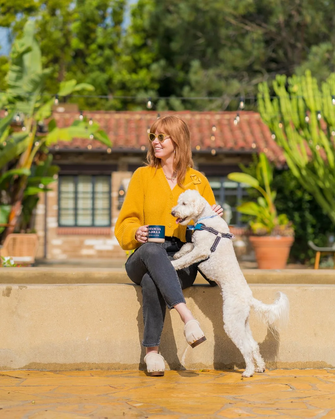 A woman with red hair, yellow sweater, and sunglasses sitting on a ledge with a white furry dog. She is holding a mug and smiling, surrounded by green plants and a brick building with string lights in the background.