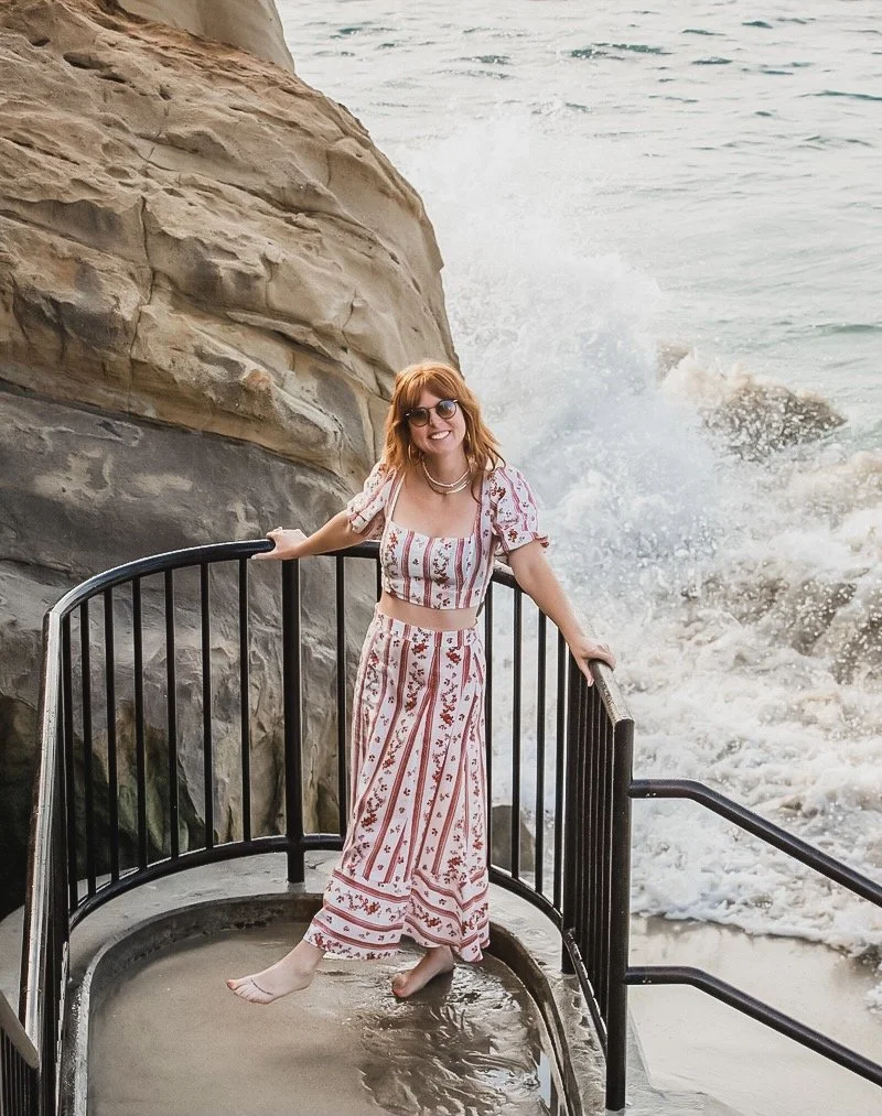 A woman with red hair wearing sunglasses, a coordinated patterned crop top and skirt, standing barefoot on a small platform near the ocean, smiling at the camera.