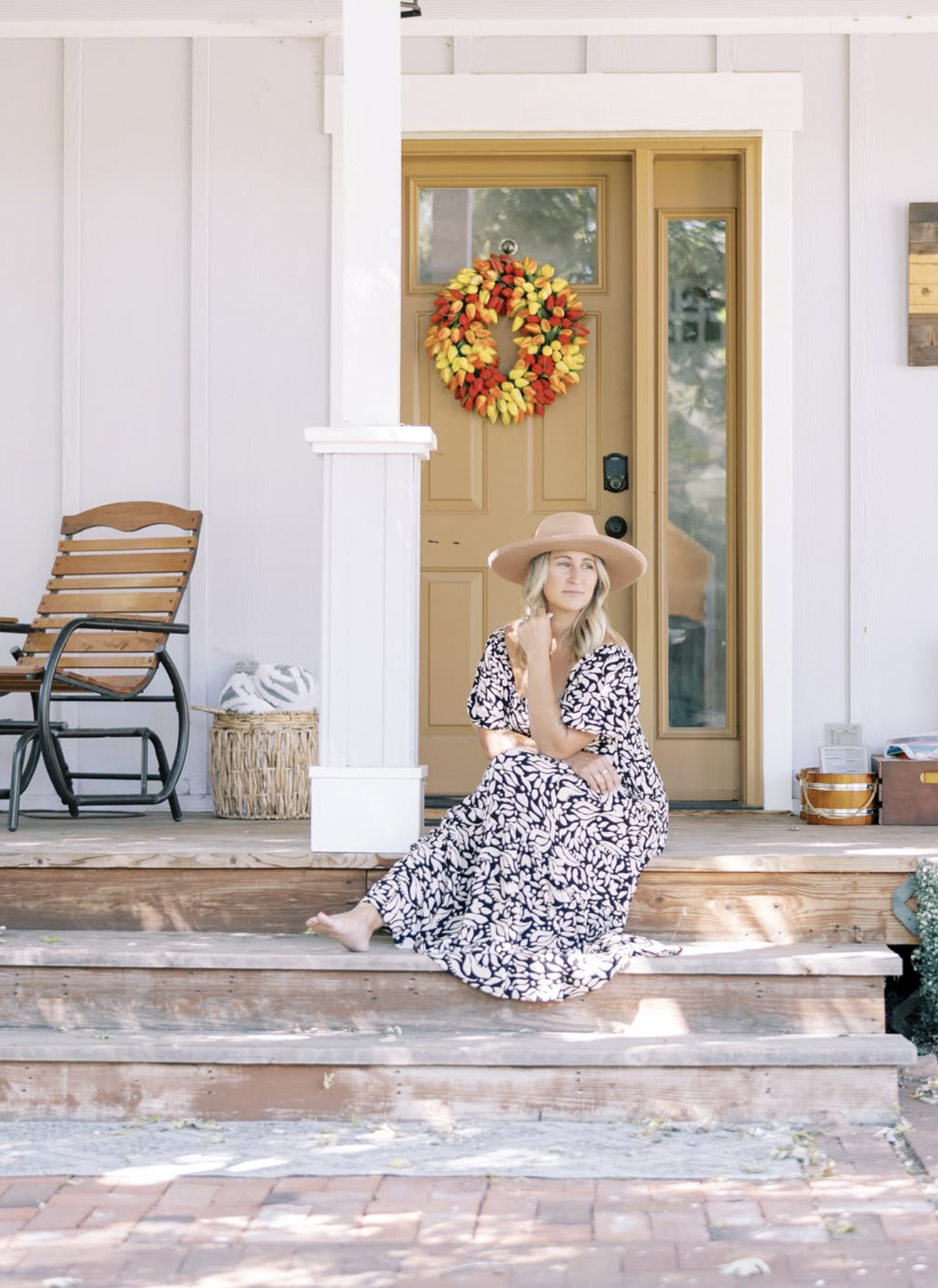 A woman in a patterned dress and wide-brimmed hat sitting on front steps of a house with a yellow door decorated with a fall wreath. There is a rocking chair, a basket, and some outdoor decor around her.