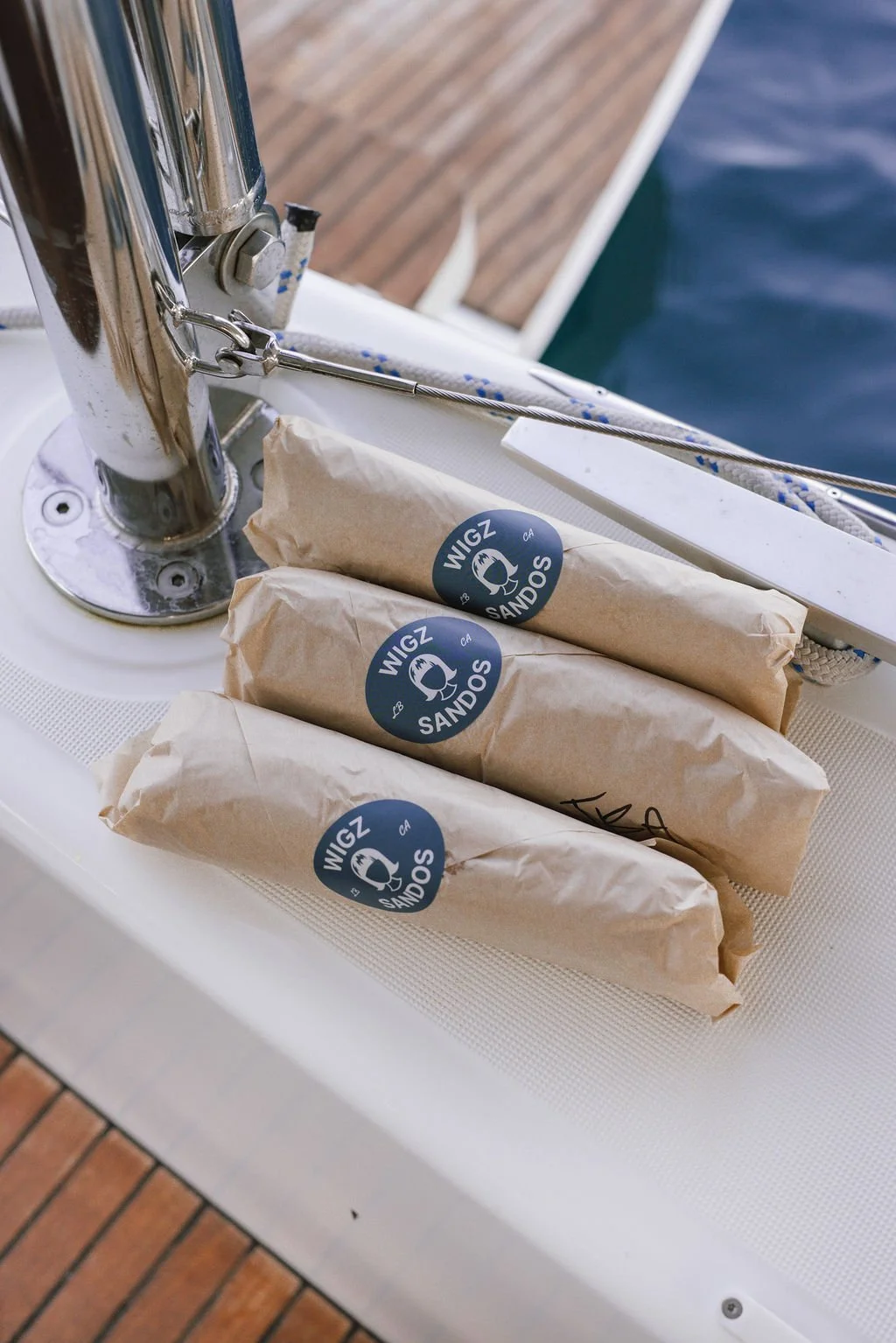 Three wrapped sandwiches labeled 'WIGZ SANDOS' on a boat deck near the water.