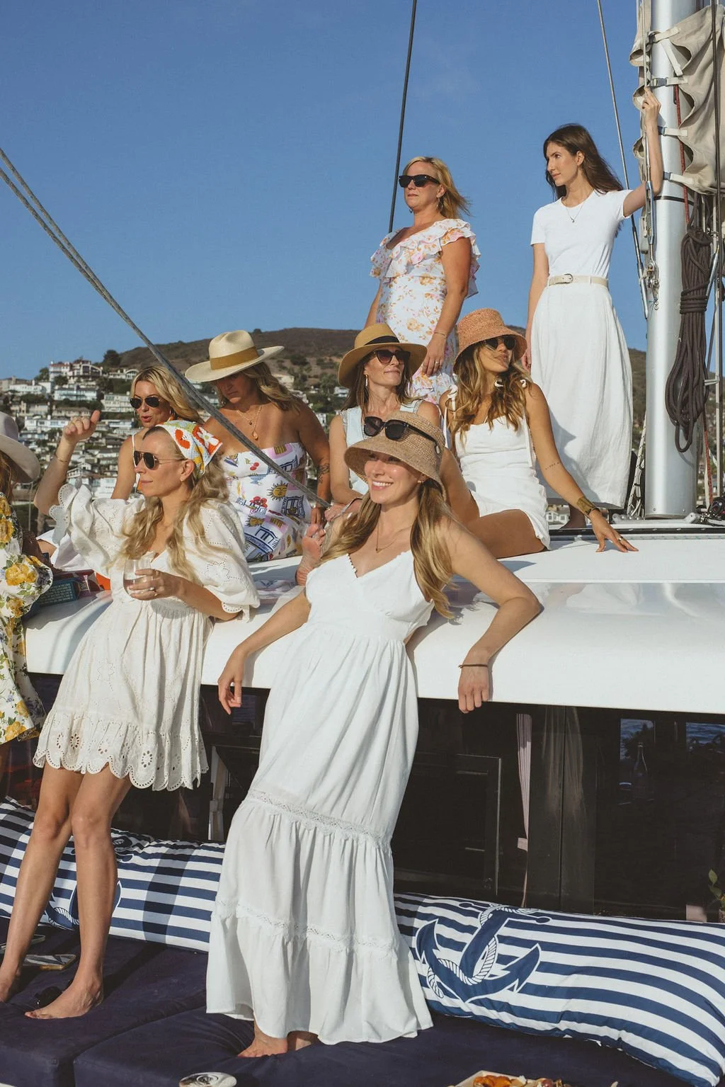A group of women wearing summer dresses and wide-brimmed hats on a yacht, with a hillside cityscape in the background.