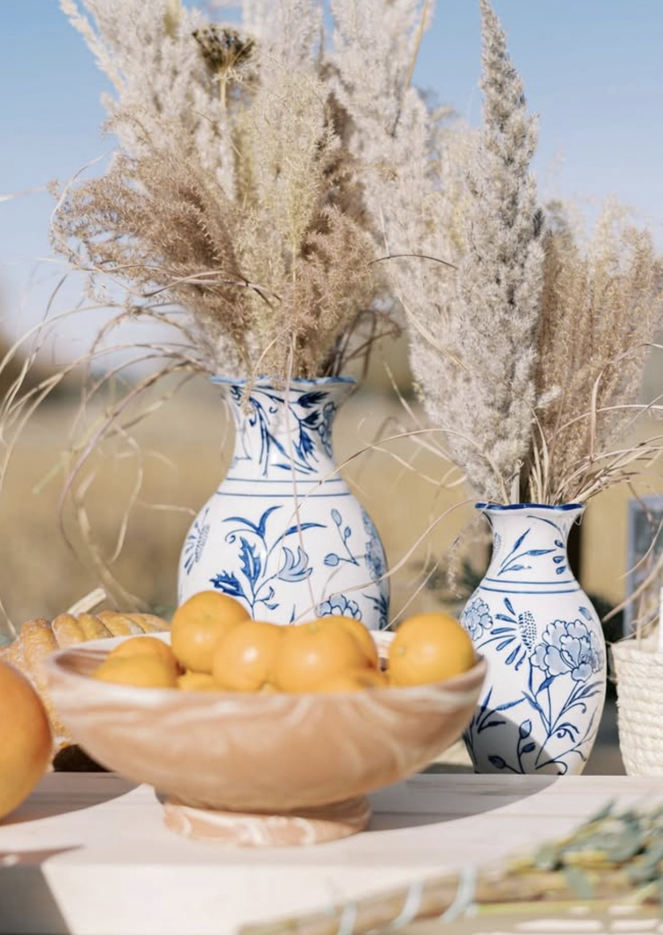 Decorative scene with two blue and white floral vases filled with dried pampas grass, a bowl of yellow oranges, and a loaf of bread on a light-colored surface.