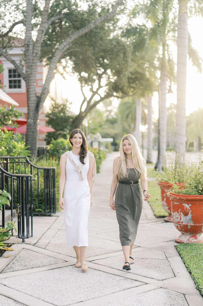 Two women walking on a sidewalk surrounded by greenery and large potted plants in a sunny outdoor setting.