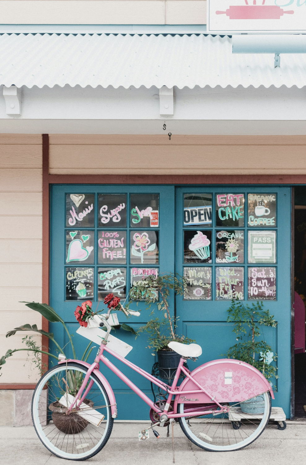 Pink bicycle parked outside a store with a blue door and windows decorated with colorful handwritten signs advertising baked goods, coffee, and organic products.
