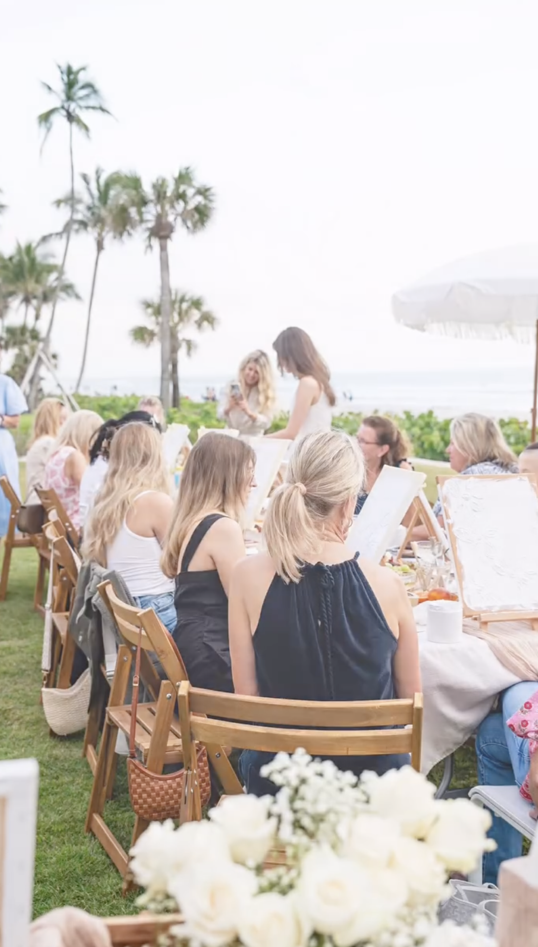 A group of women gathered outdoors at a beachside picnic, sitting on wooden chairs around a long table, with palm trees and the ocean in the background.