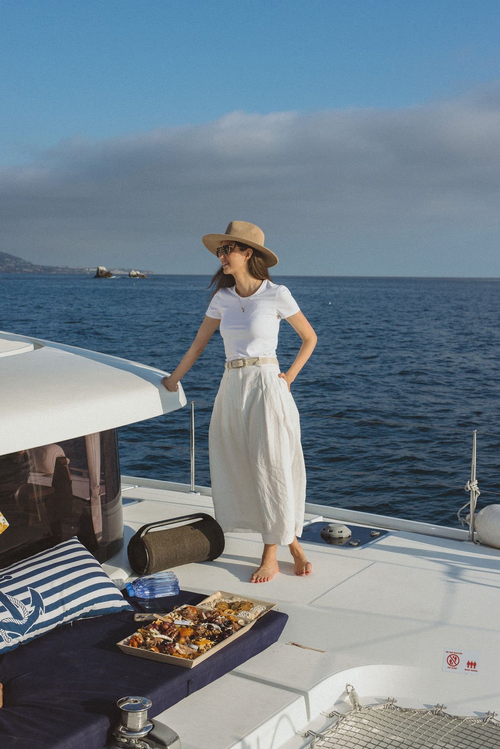 A woman wearing a white t-shirt, beige wide-leg pants, straw hat, and sunglasses standing on a boat with a tray of food, the ocean in the background.