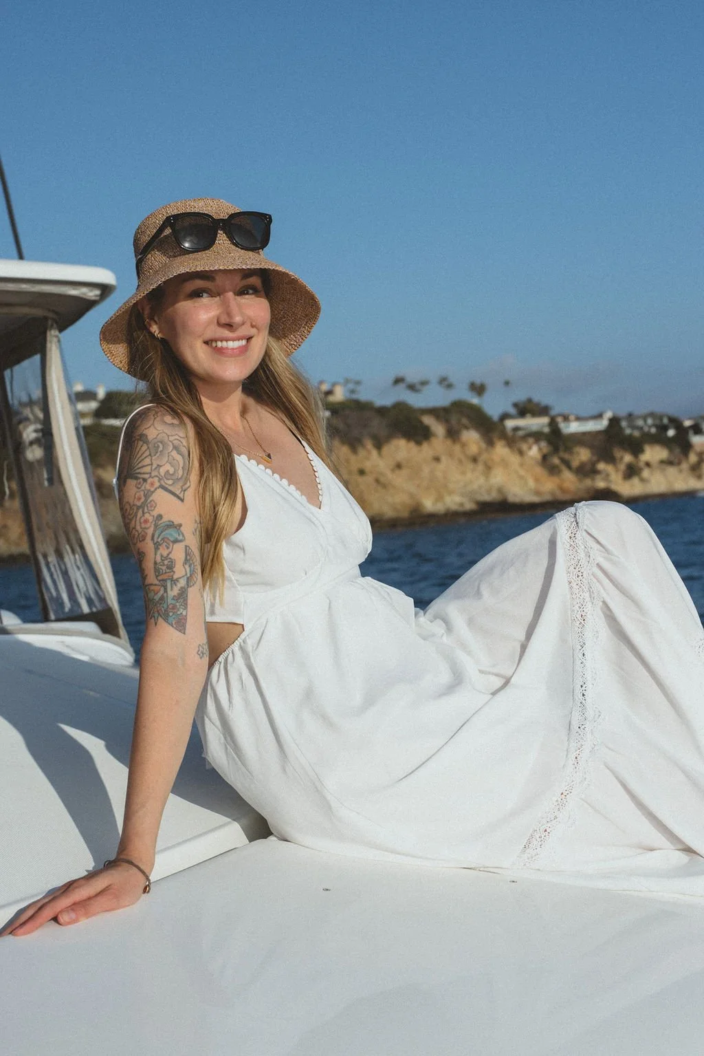 A woman with tattoos wearing a white dress and a straw hat with sunglasses sitting on a boat, smiling, with a view of the ocean and cliffs in the background.