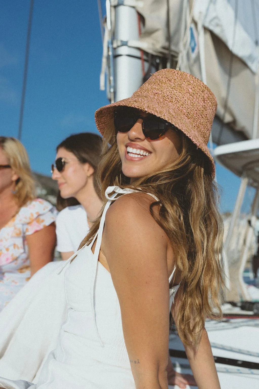 A smiling woman with long wavy hair wearing a pink hat and black sunglasses, standing outdoors near a boat with other women in the background.