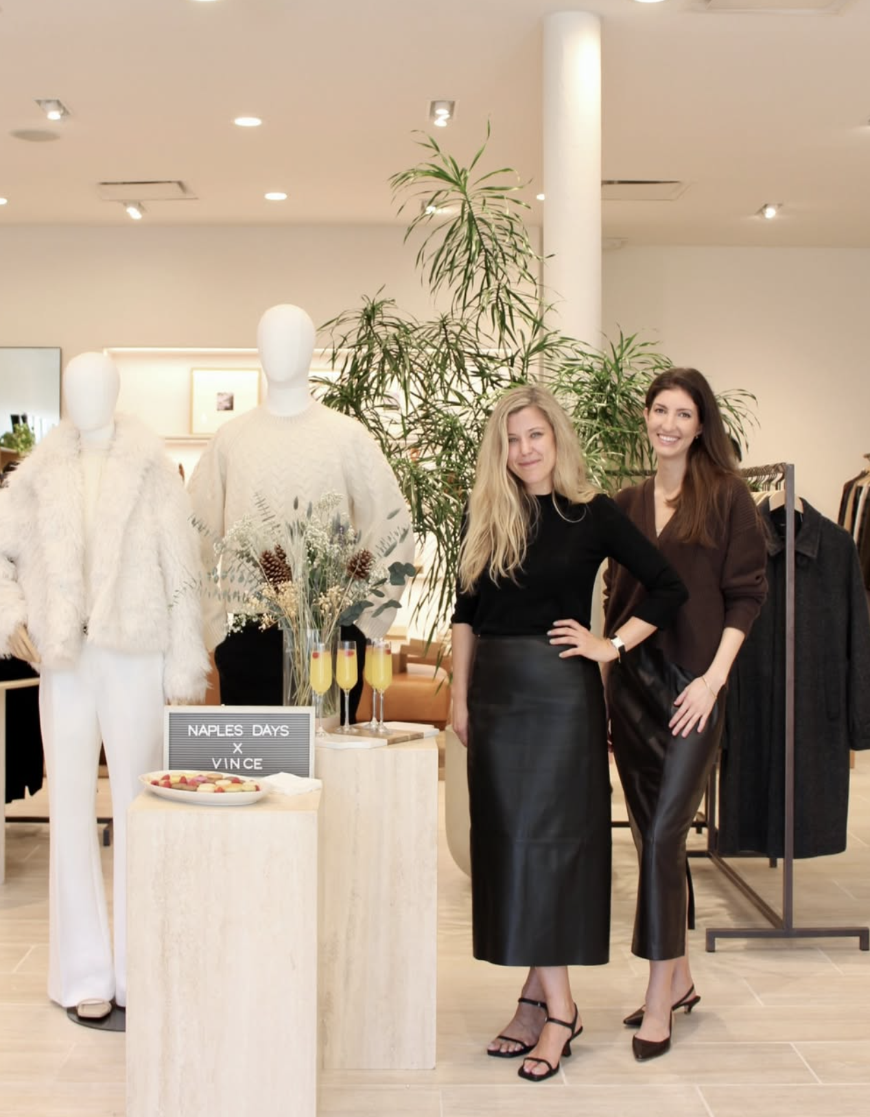Two women standing inside a clothing store with mannequins dressed in fur and knit sweaters and clothing racks in the background. There is a floral arrangement and drinks on a small table.