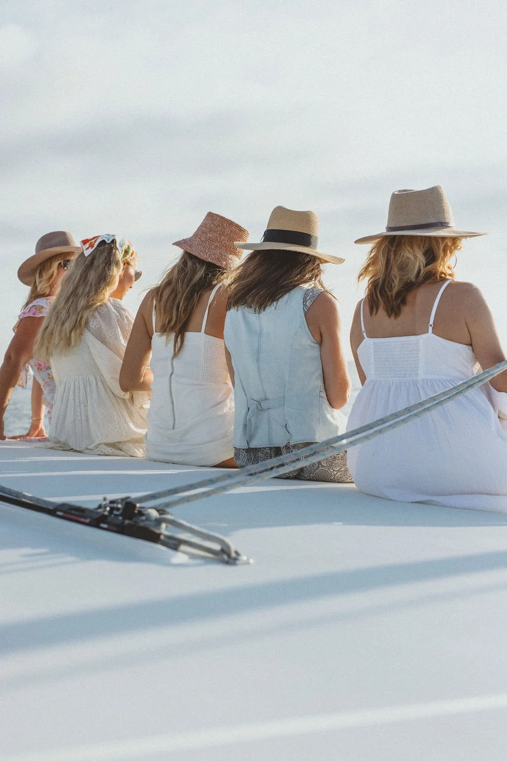 Group of women sitting on a boat deck, looking at the ocean, wearing summer dresses and wide-brimmed hats, enjoying a sunny day.