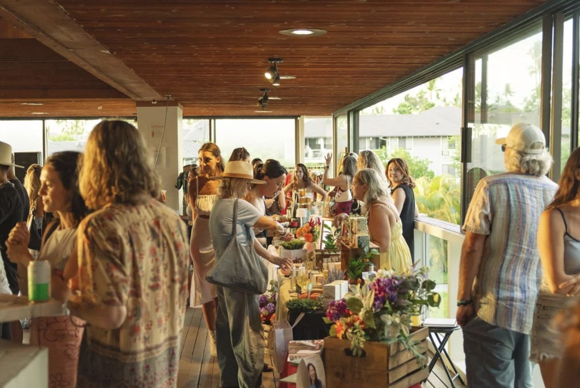 Women and men browsing and shopping at a flower and gift market inside a store with large windows, wooden ceiling, and natural light.