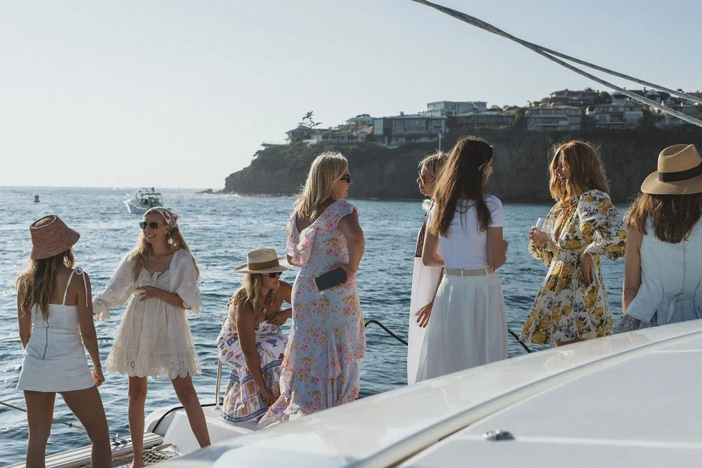Group of women in summer dresses and hats conversing on a yacht near the coast with houses on cliffs in the background.
