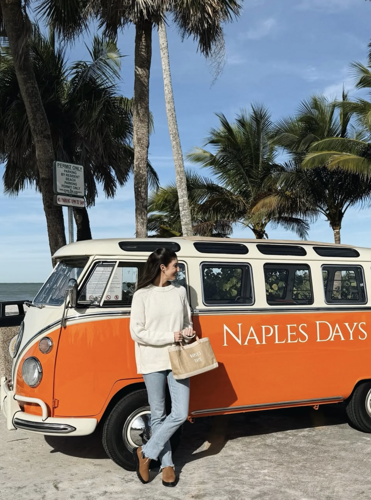 A woman standing next to an orange and white vintage van with sign reading 'Naples Days' parked on a beach with palm trees and a cloudy sky in the background.