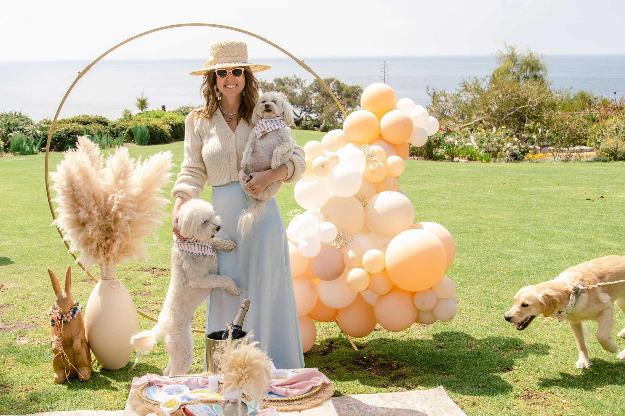 A woman in a beige cardigan, blue skirt, wearing sunglasses and a wide-brimmed hat, smiling and holding two small dogs dressed with bandanas, standing outdoors on a grassy area with balloons and decorative elements.