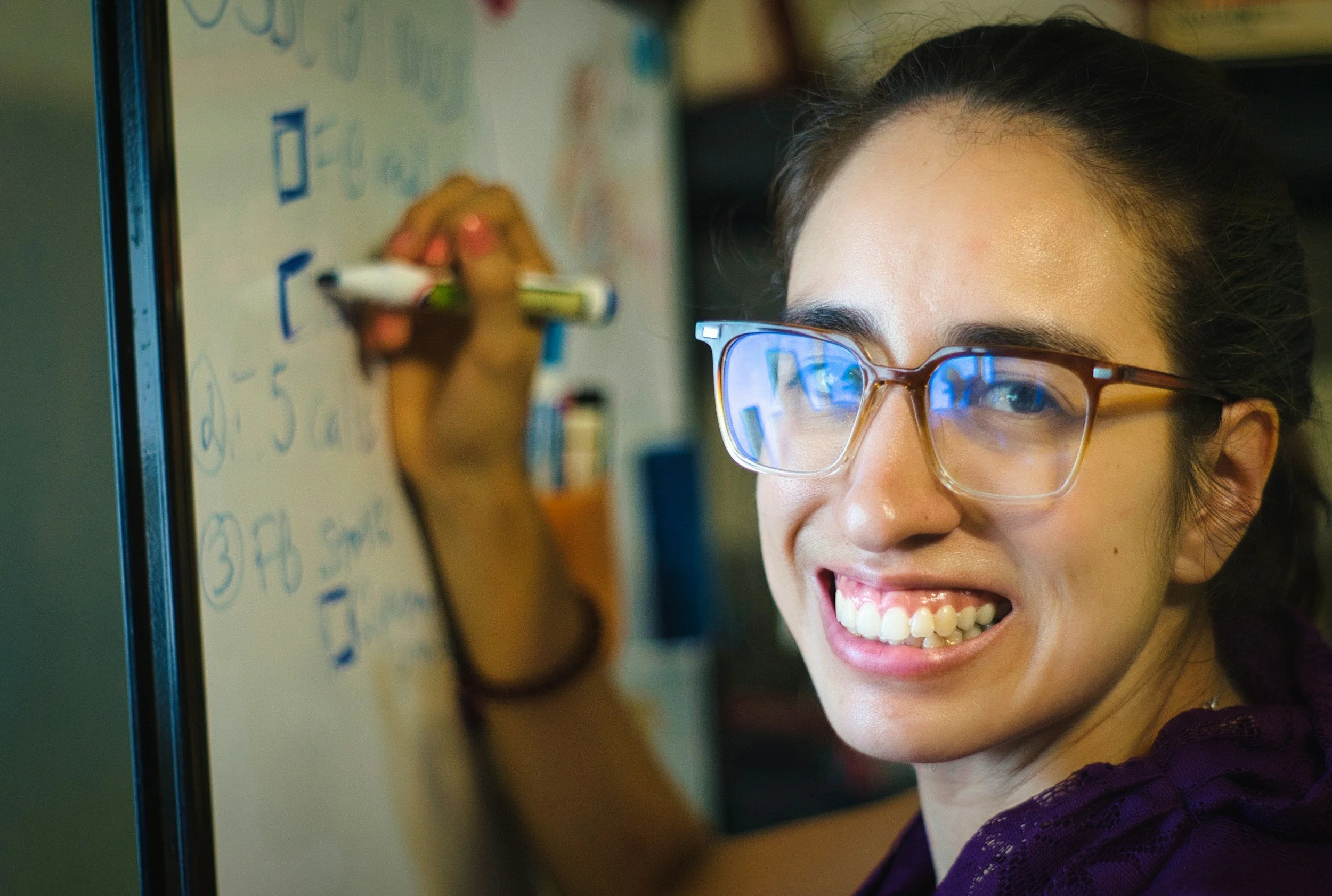 Close-up of a young woman with glasses smiling as she writes math problems on a whiteboard in a classroom.