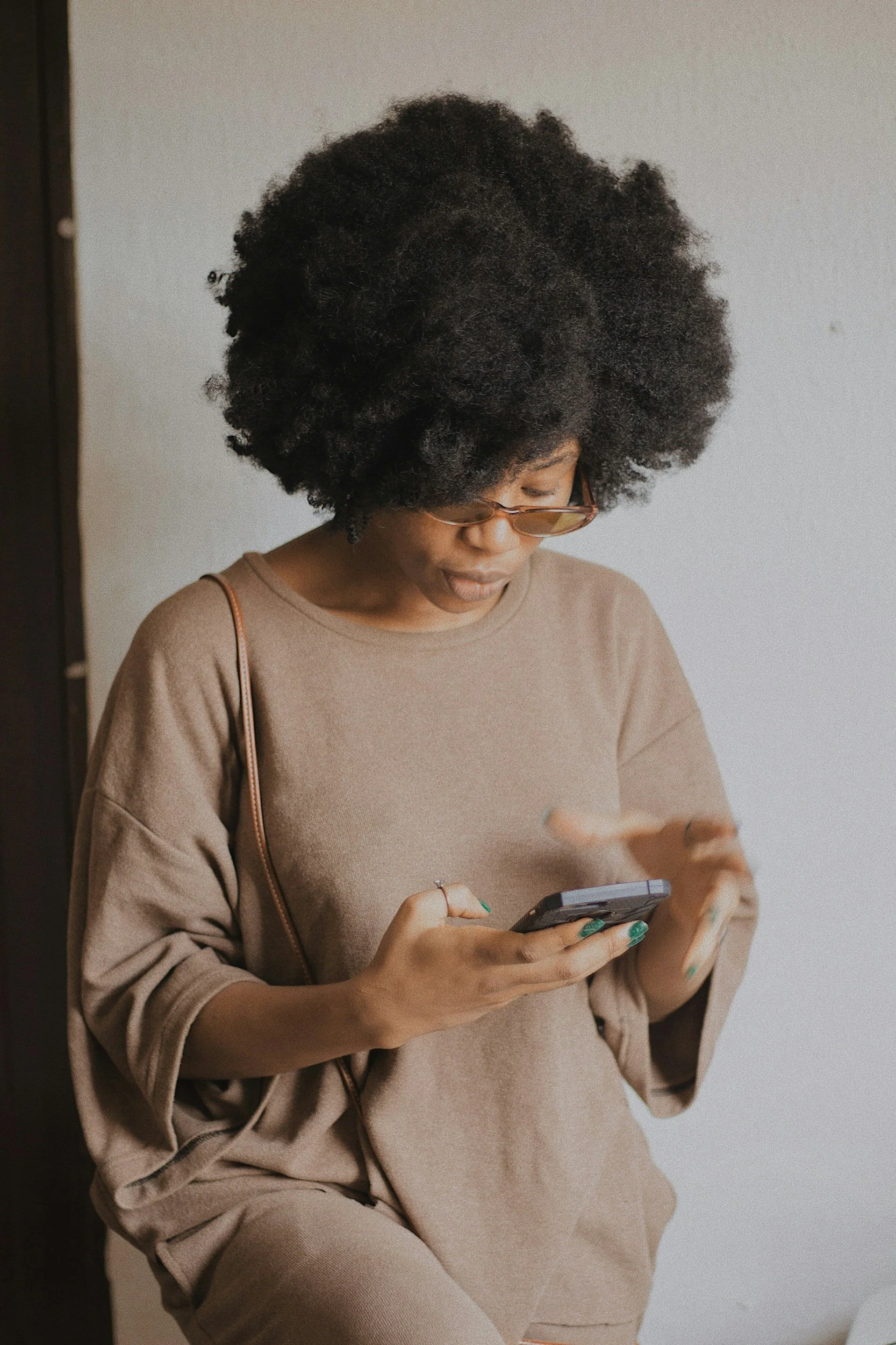 A woman with natural curly hair, wearing glasses and a beige outfit, is looking at her smartphone.