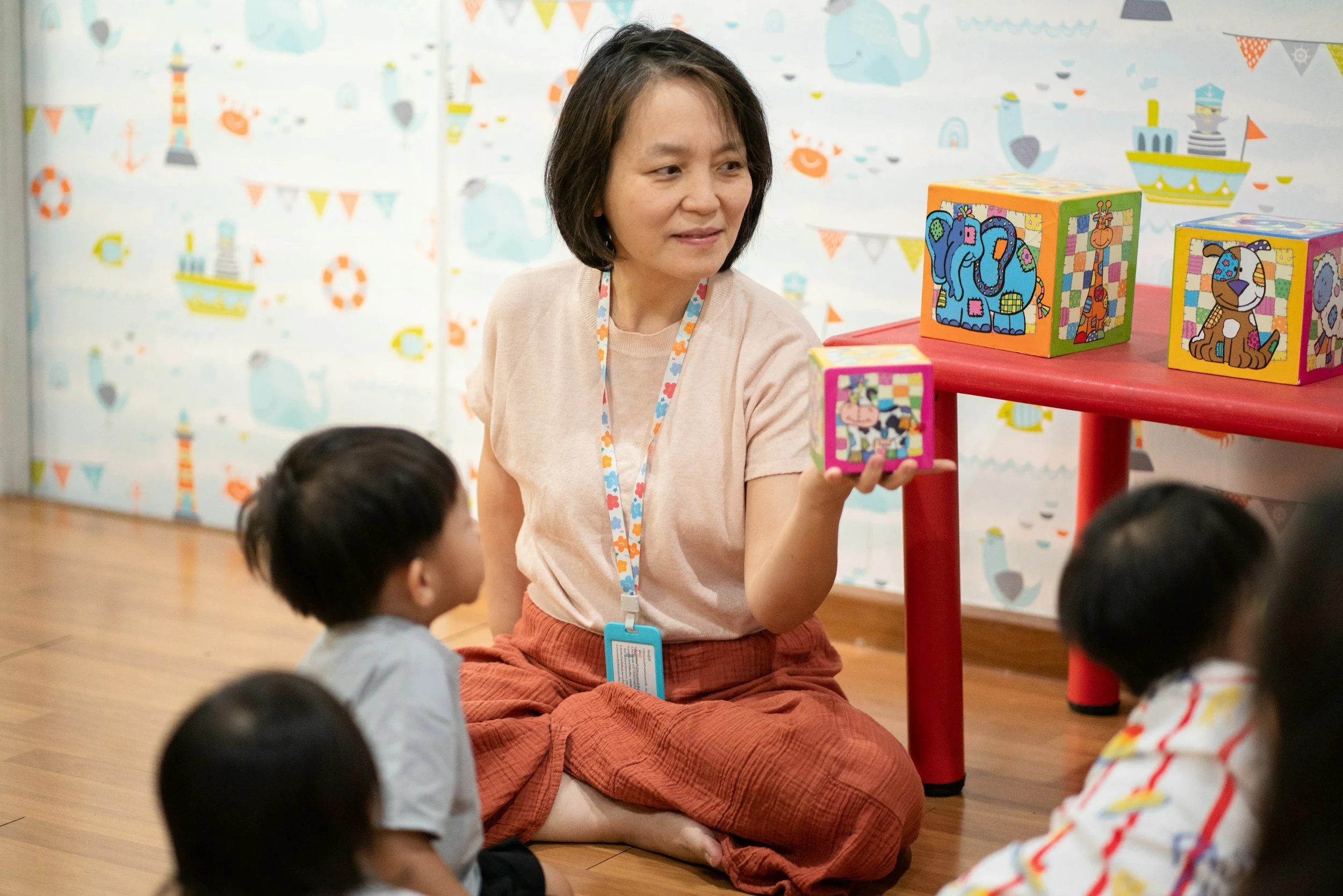 A woman with short dark hair, wearing a pink top, orange skirt, and a lanyard, is sitting on the floor, engaging with young children in a colorful playroom. She holds a small block with an animal illustration, and there are decorated blocks with animal images on a red table nearby. The background features a bright, playful wall with various maritime-themed illustrations such as ships, anchors, and life preservers.