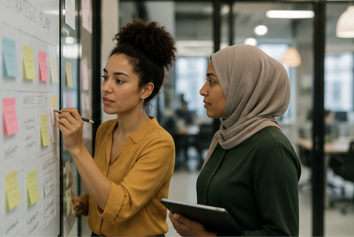 Two women in an office, one writing on a whiteboard filled with sticky notes, the other observing and holding a tablet.
