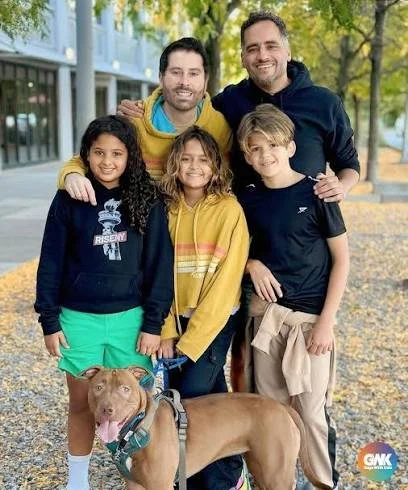 Group of four children with two adult men, standing outdoors in fall with a dog, smiling at the camera.