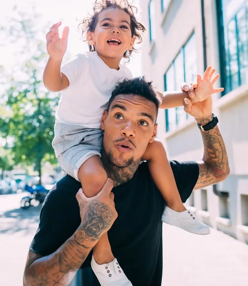 A young girl with curly hair riding on a man's shoulders, outside on a sunny day, both smiling and appearing joyful.