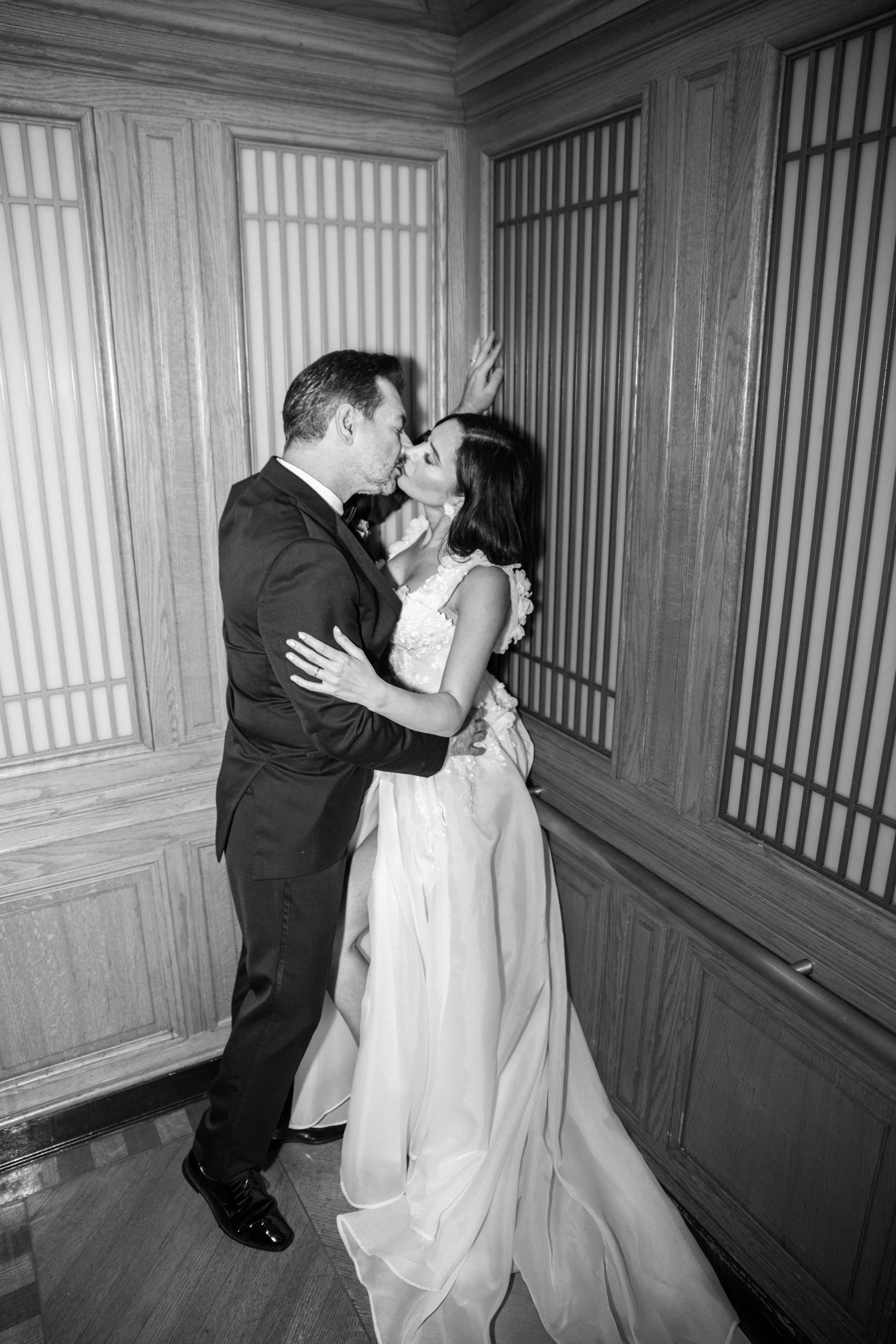 A black-and-white photo of a couple in wedding attire sharing a kiss in a room with wooden paneling and traditional screen doors.
