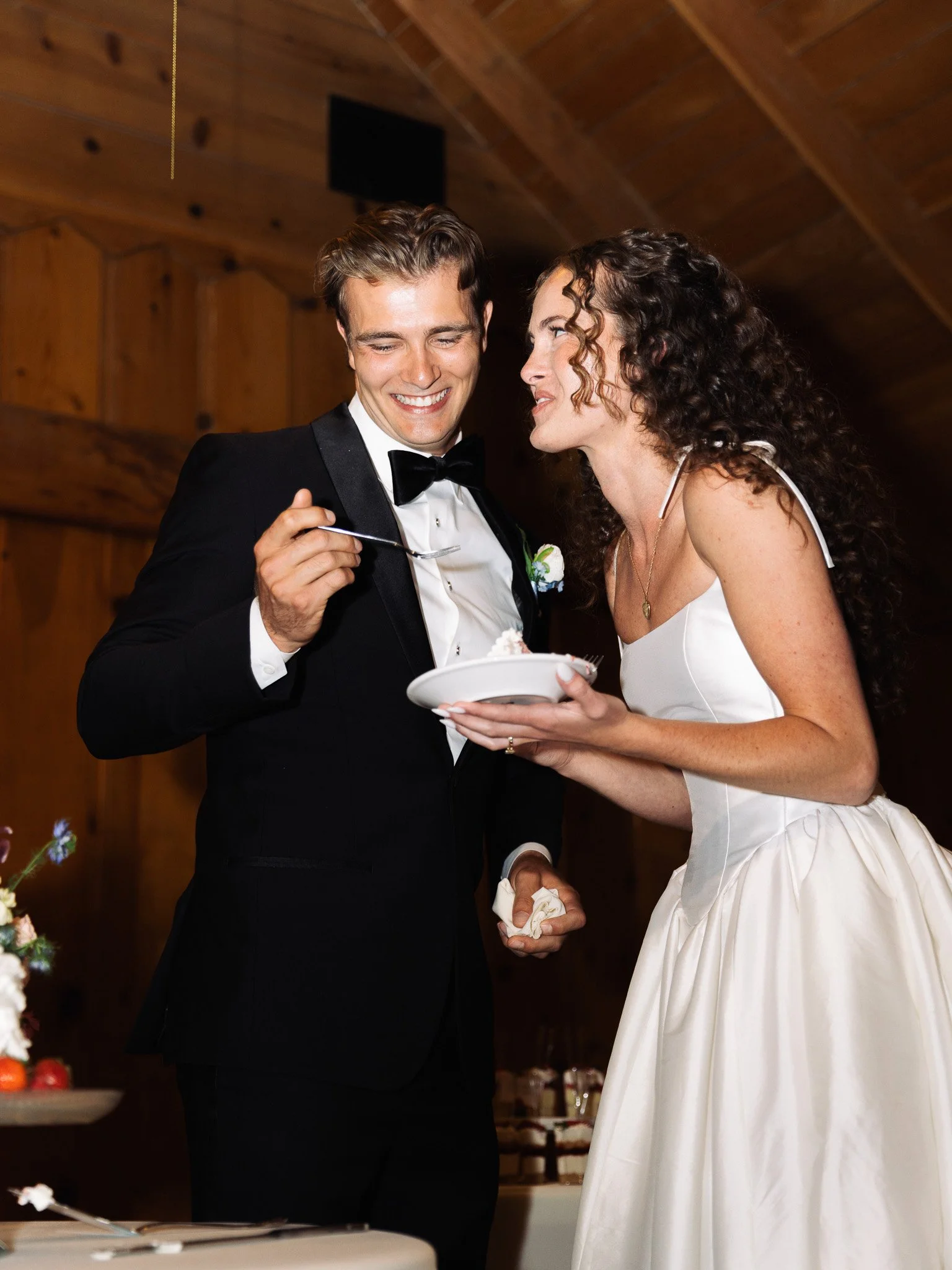 A newlywed couple sharing wedding cake, the groom in a tuxedo and the bride in a white wedding dress, indoors with wooden walls.