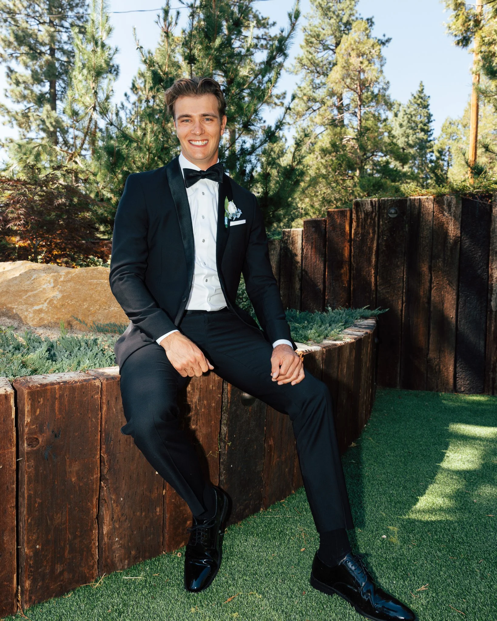 A man in a black tuxedo and bow tie sitting on a wooden ledge outdoors, with green trees and blue sky in the background.