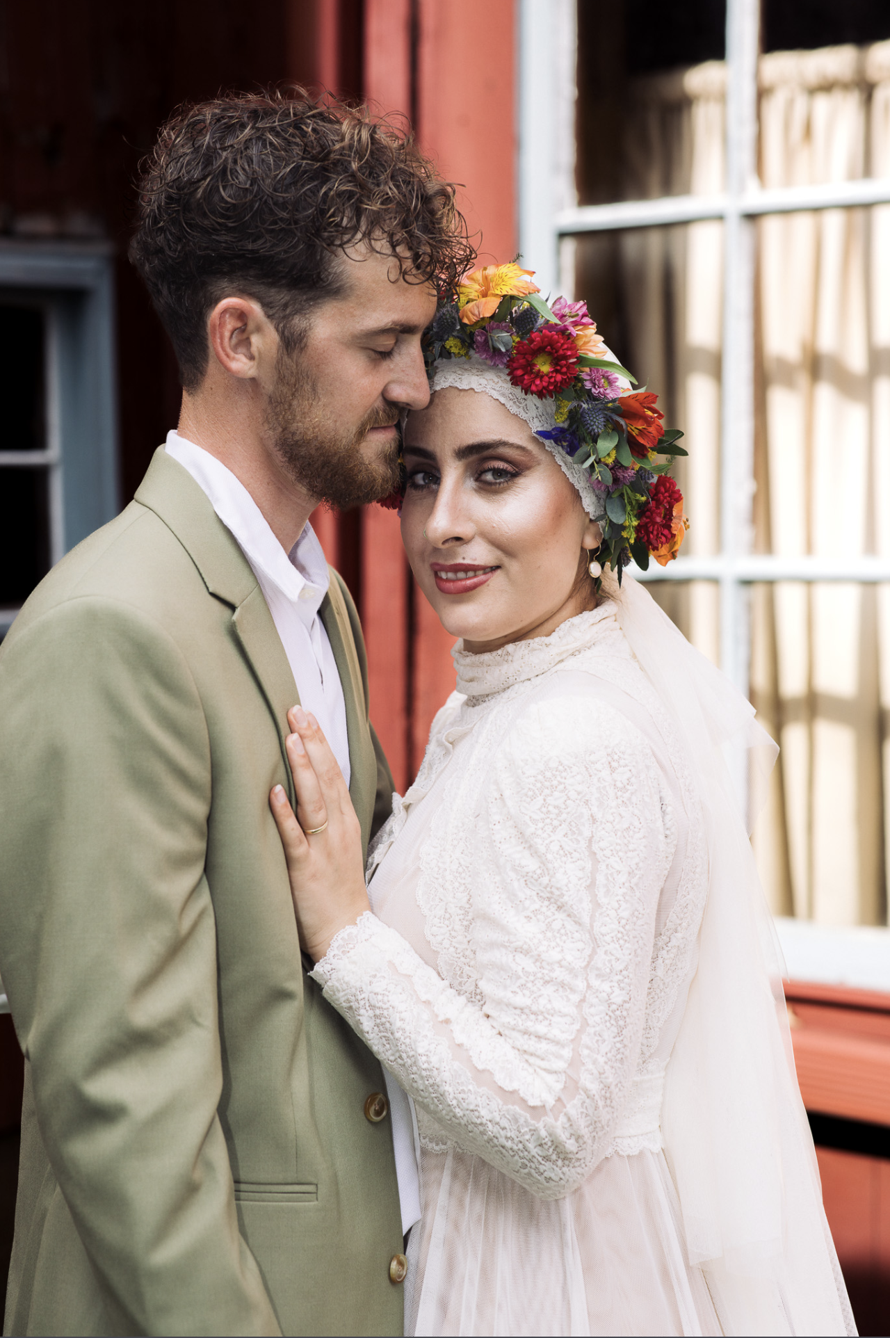A bride and groom stand close together, with the groom resting his forehead against the bride's forehead. The bride is wearing a white lace wedding dress and a colorful flower crown, while the groom is dressed in a light green suit and white shirt, w