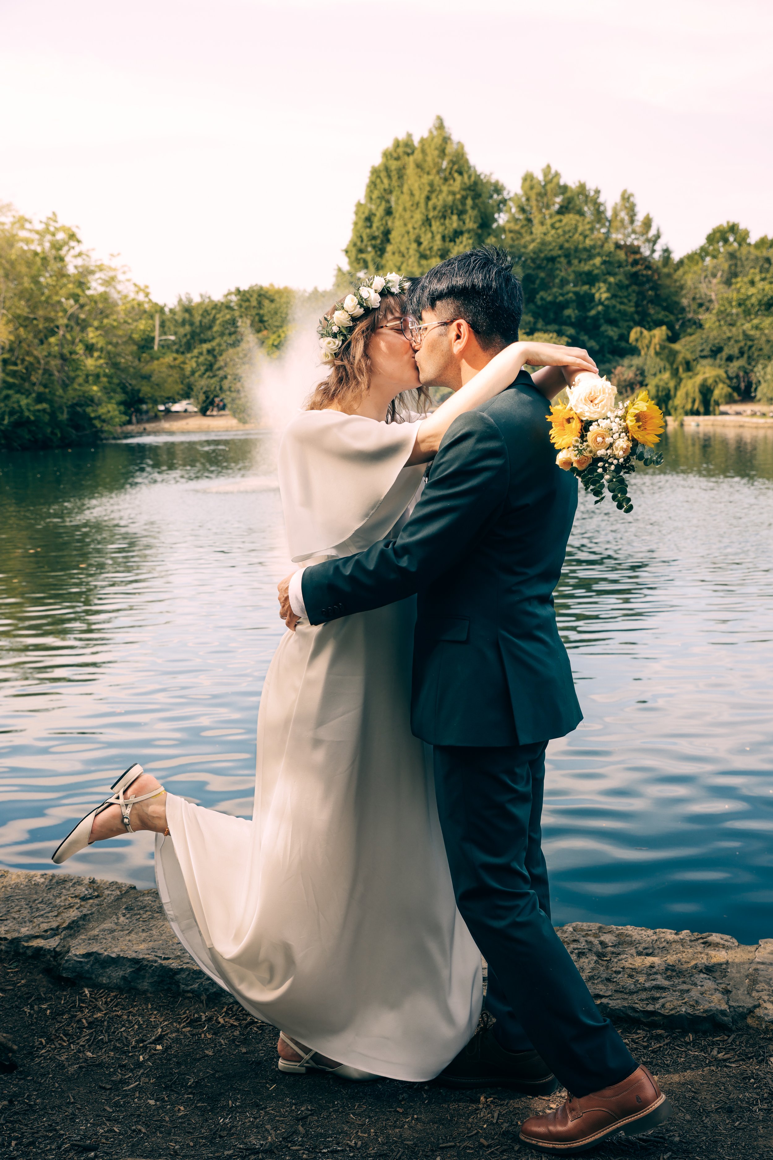 A couple in wedding attire sharing a kiss by a lake, with the woman wearing a flower crown and holding a bouquet, and the man holding her as she leans back with one foot raised.