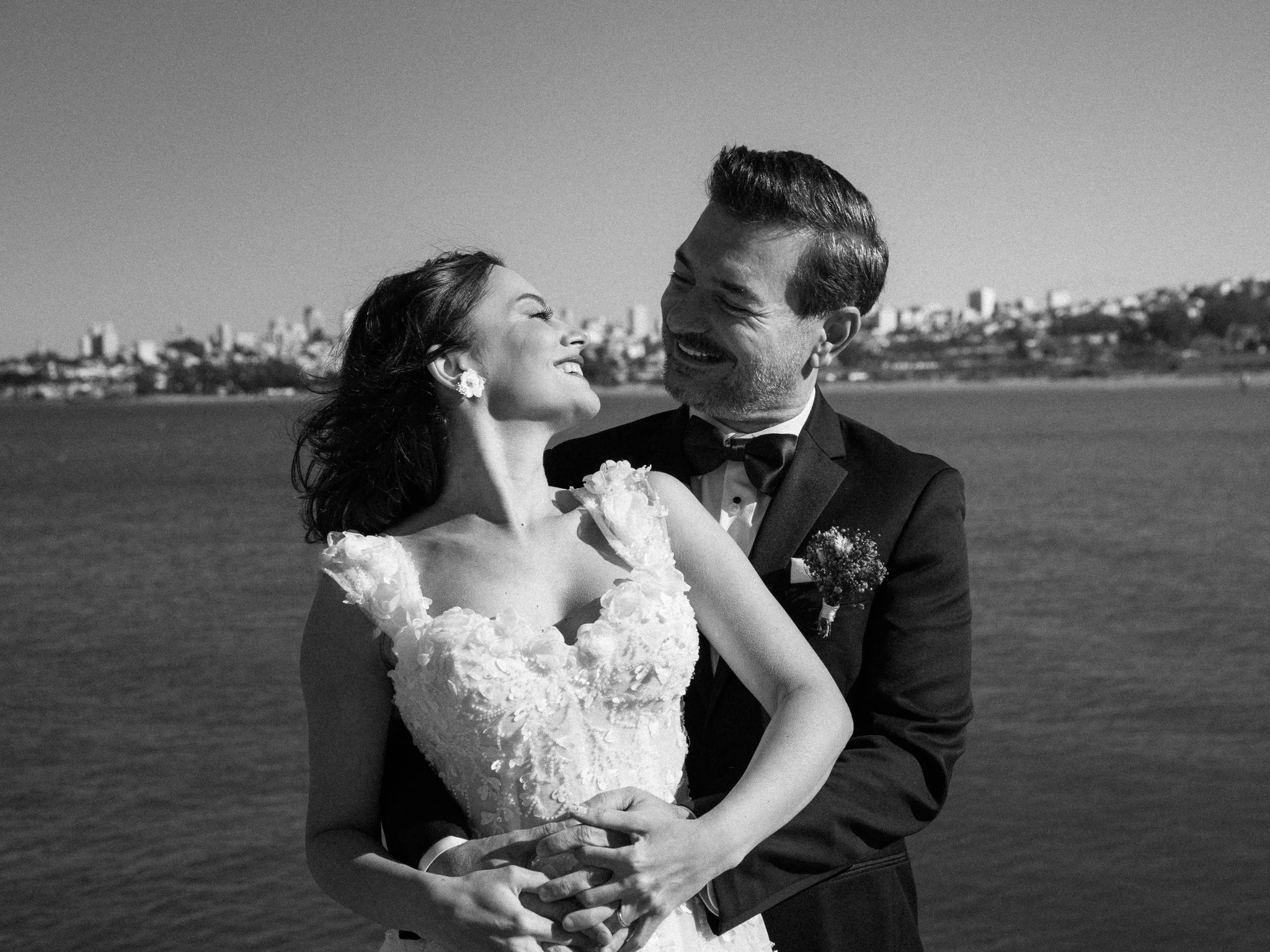 A black-and-white photo of a newlywed couple smiling and looking at each other by the water, with a city skyline in the background.