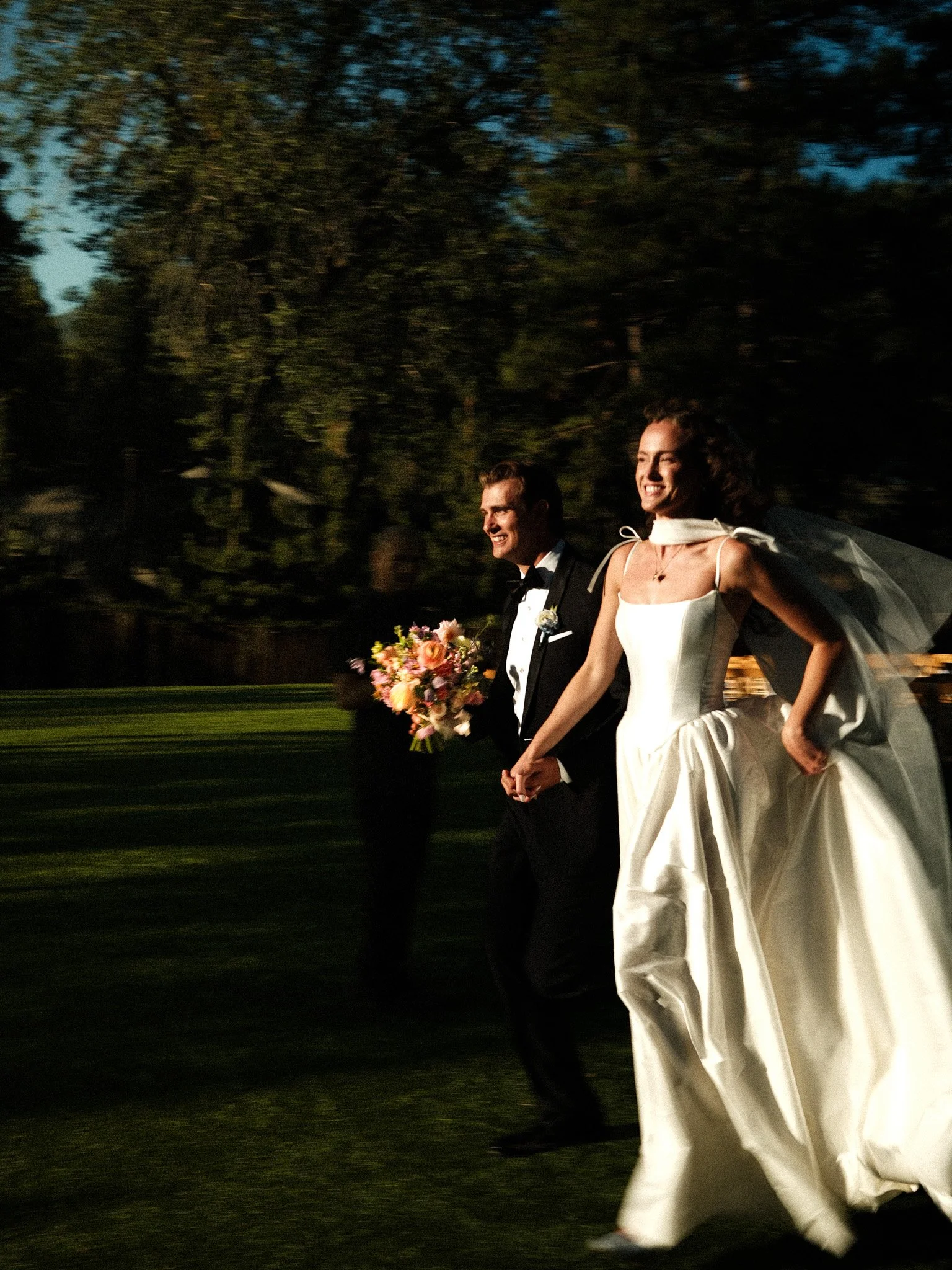 A bride and groom holding hands and smiling while walking outdoors on their wedding day in the evening, with a person carrying a bouquet of flowers in the background.