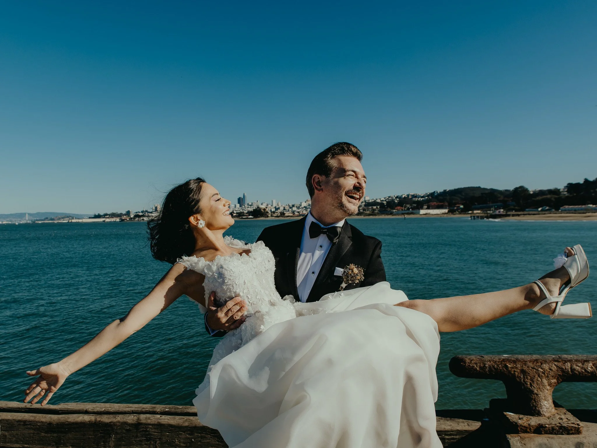 A smiling bride and groom in wedding attire sitting on a dock by the water, with the groom holding the bride in his arms as she extends her arms outward, with a city skyline in the background.