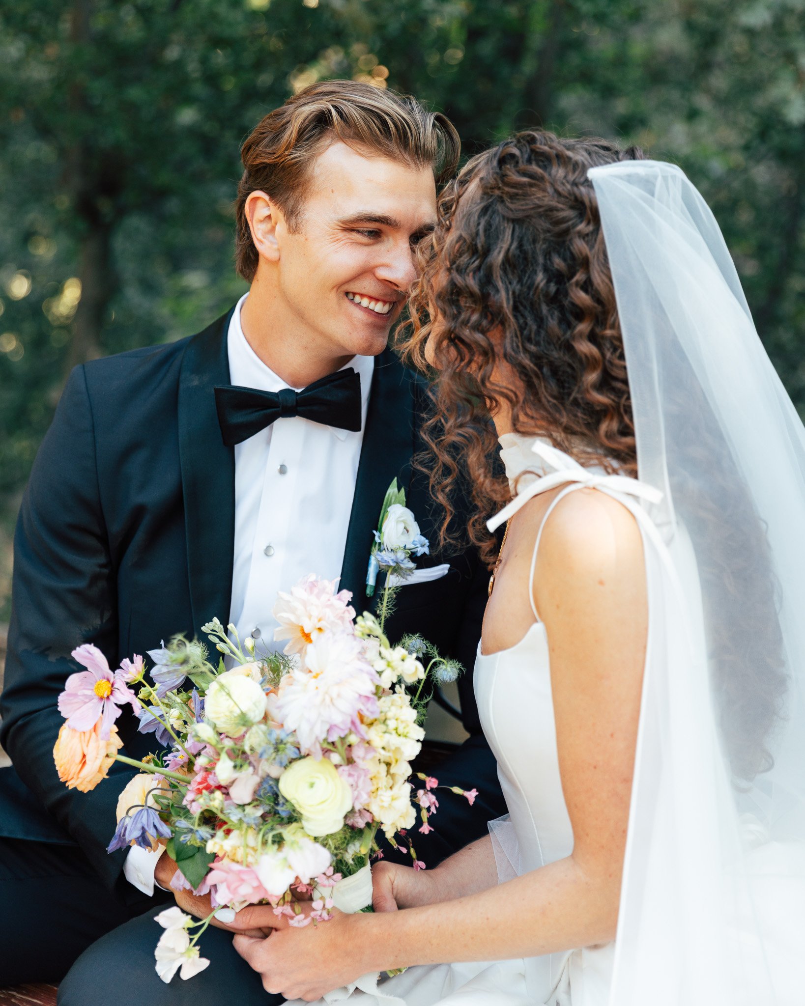 A newlywed couple smiling and gazing at each other, outdoors, in wedding attire, holding a bouquet of flowers.